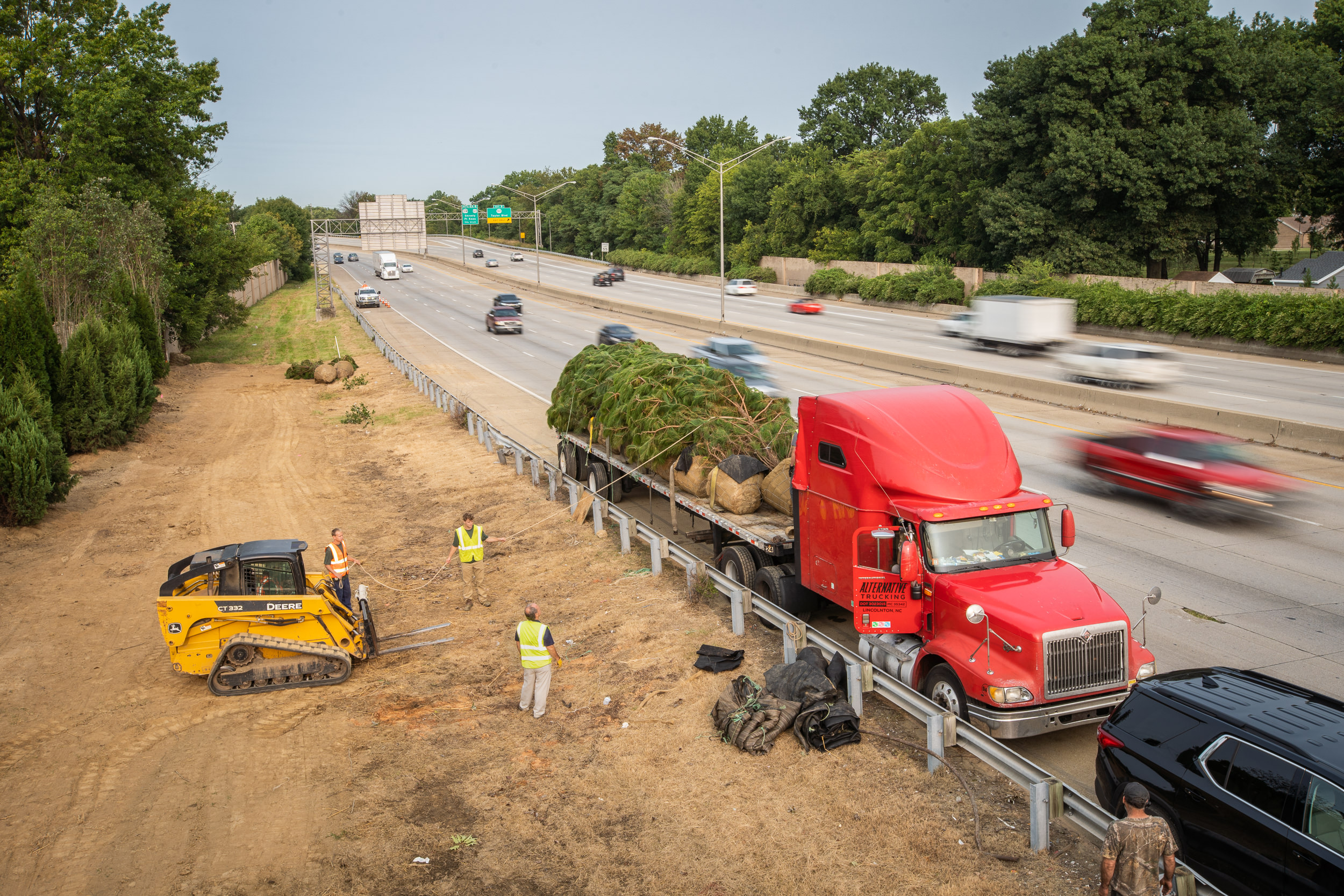 Cars speed by a flatbed semi truck parked on the shoulder of a highway, laden with mature trees ready for planting.