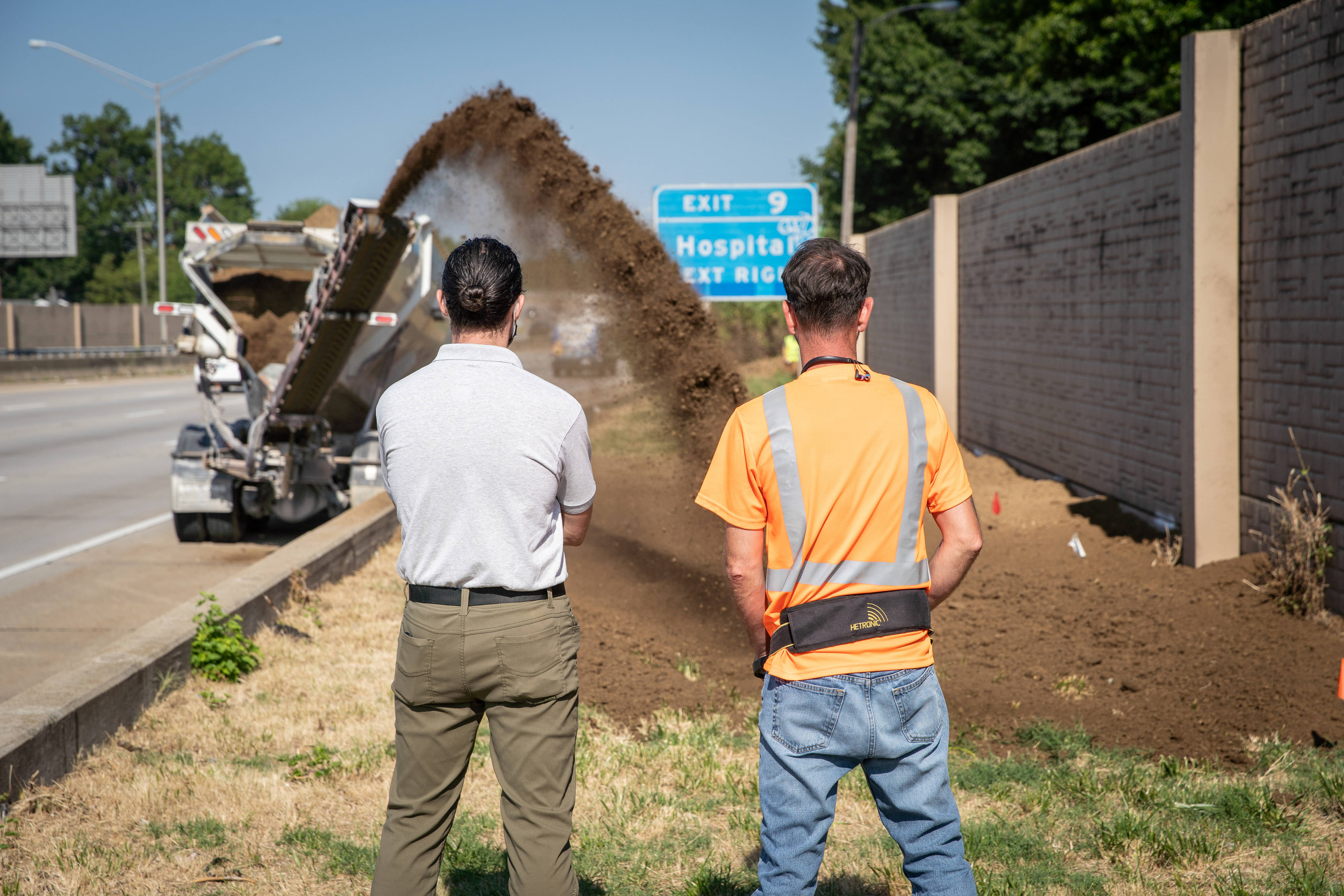 Two men stand together watching soil being spread from the back of a dump truck onto a planting area tucked between a highway and tall sound barrier wall.