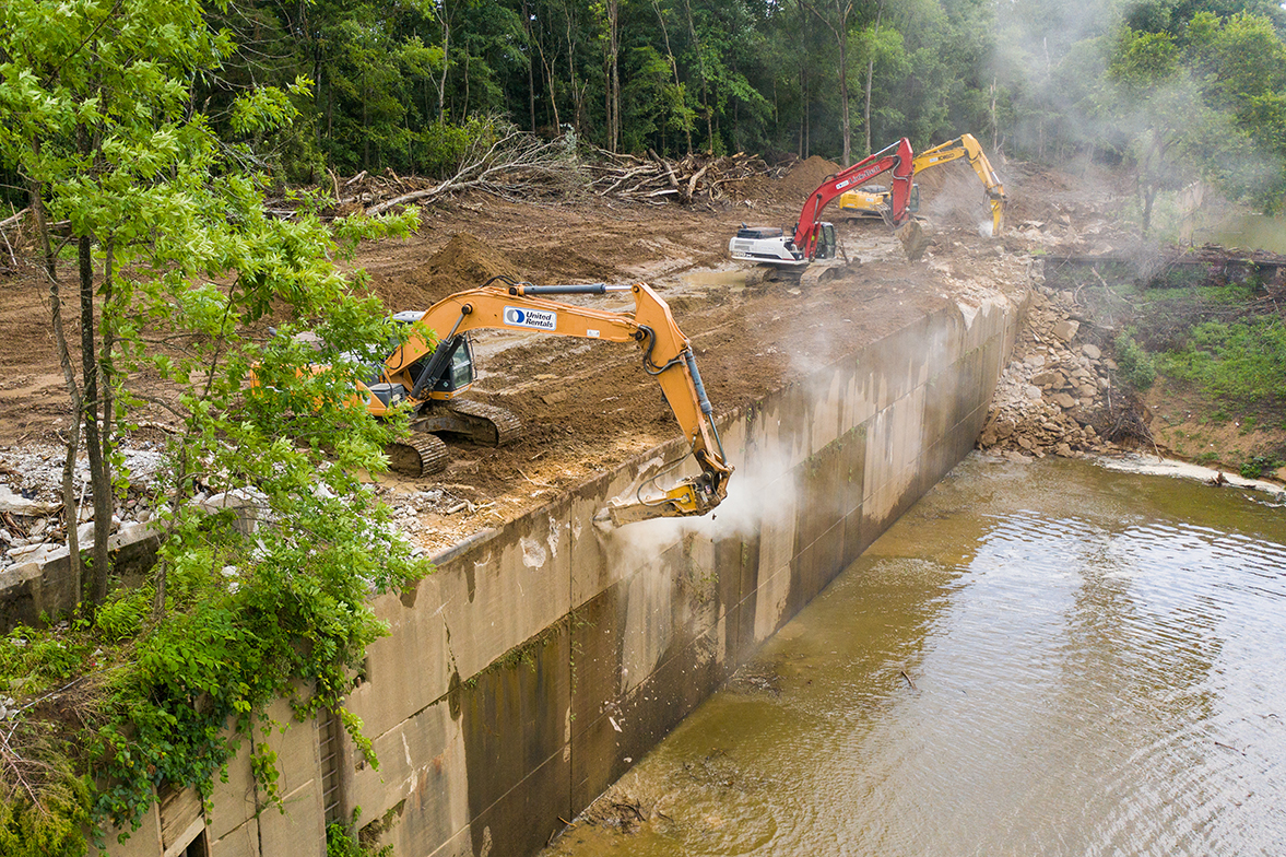 Large equipment hammers away at a large concrete structure on a river.