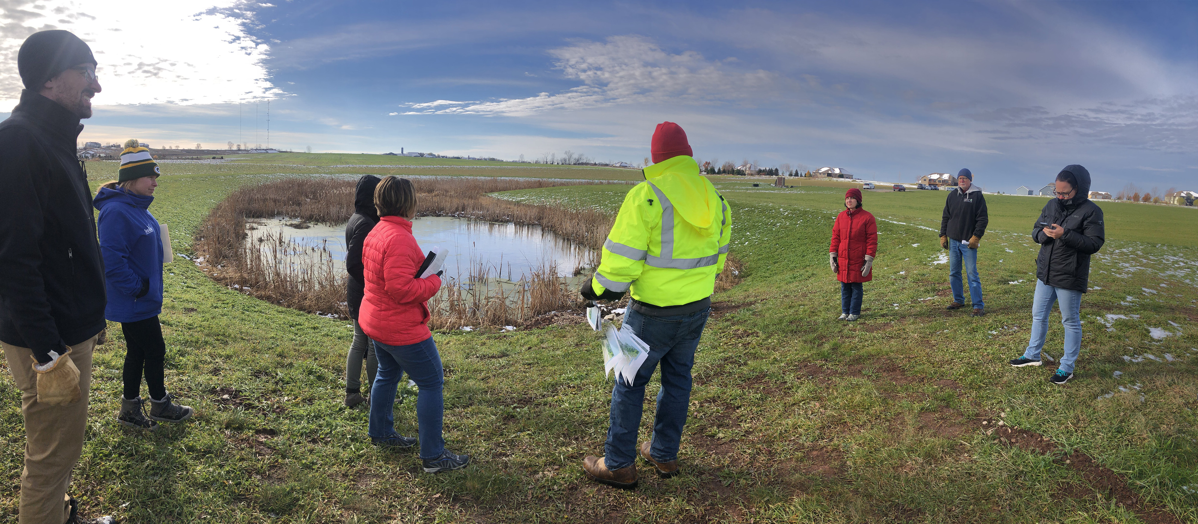 Eight people dressed in winter clothing stand around a small pond surrounded by cattails and agricultural land with a blue sky overhead.