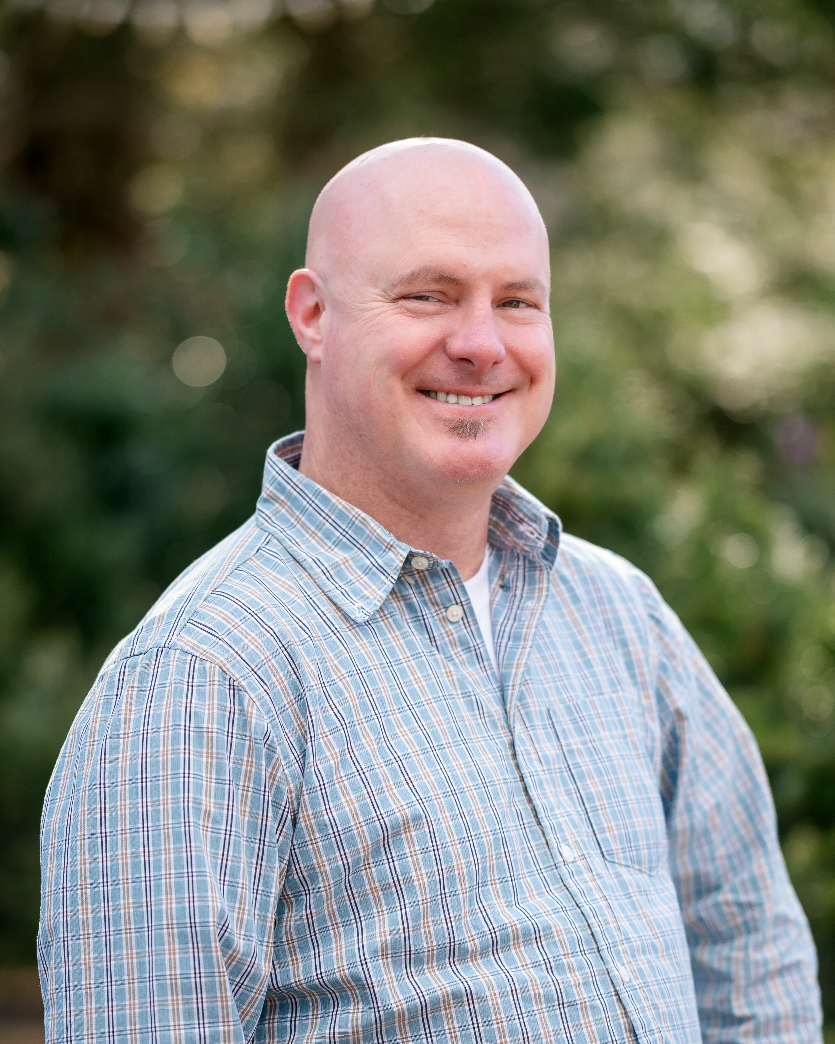 Headshot of Rene Wendell in a blue plaid shirt with greenery in the background. 