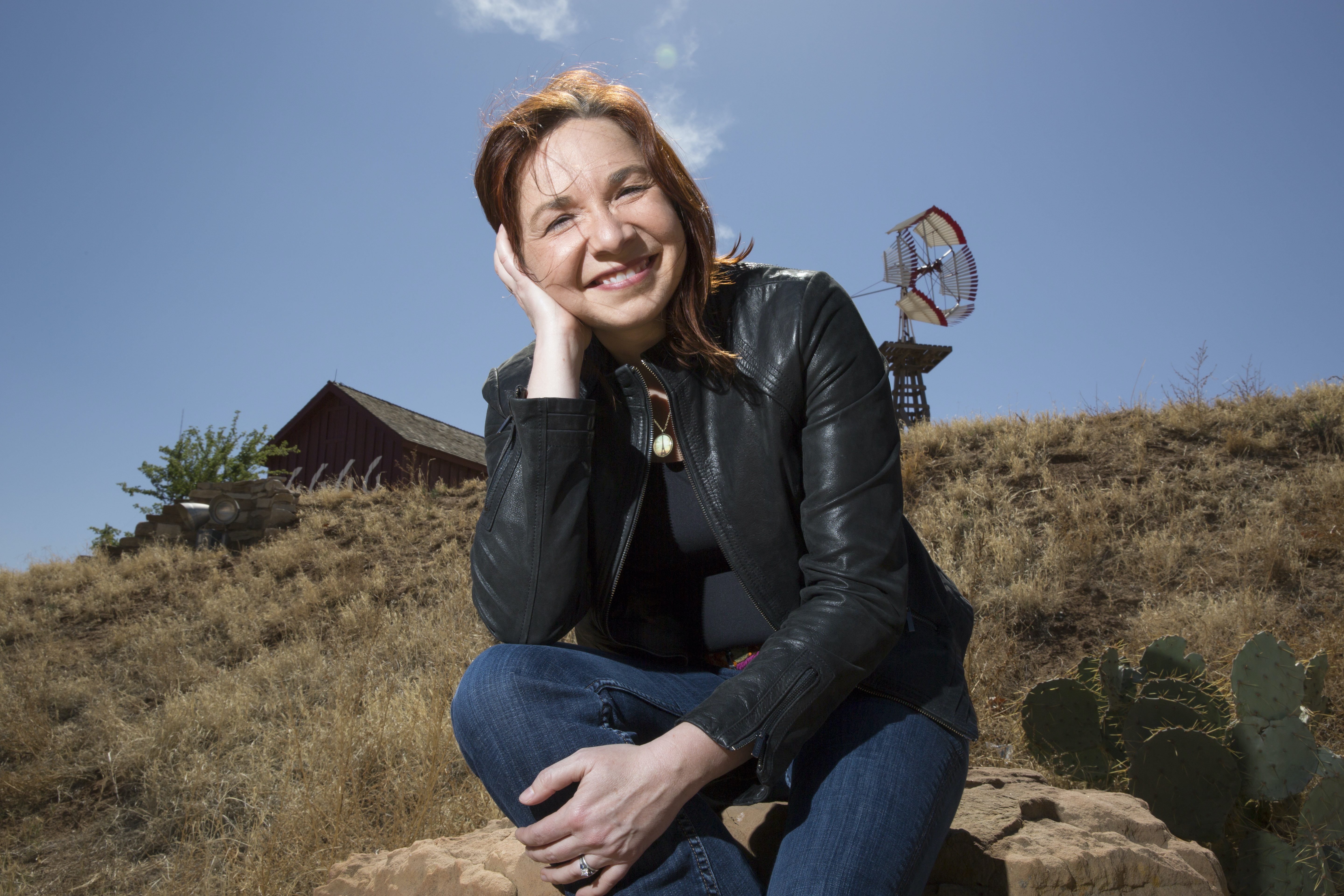 A photo of a woman kneeling outside in grass smiling with a barn in the background.