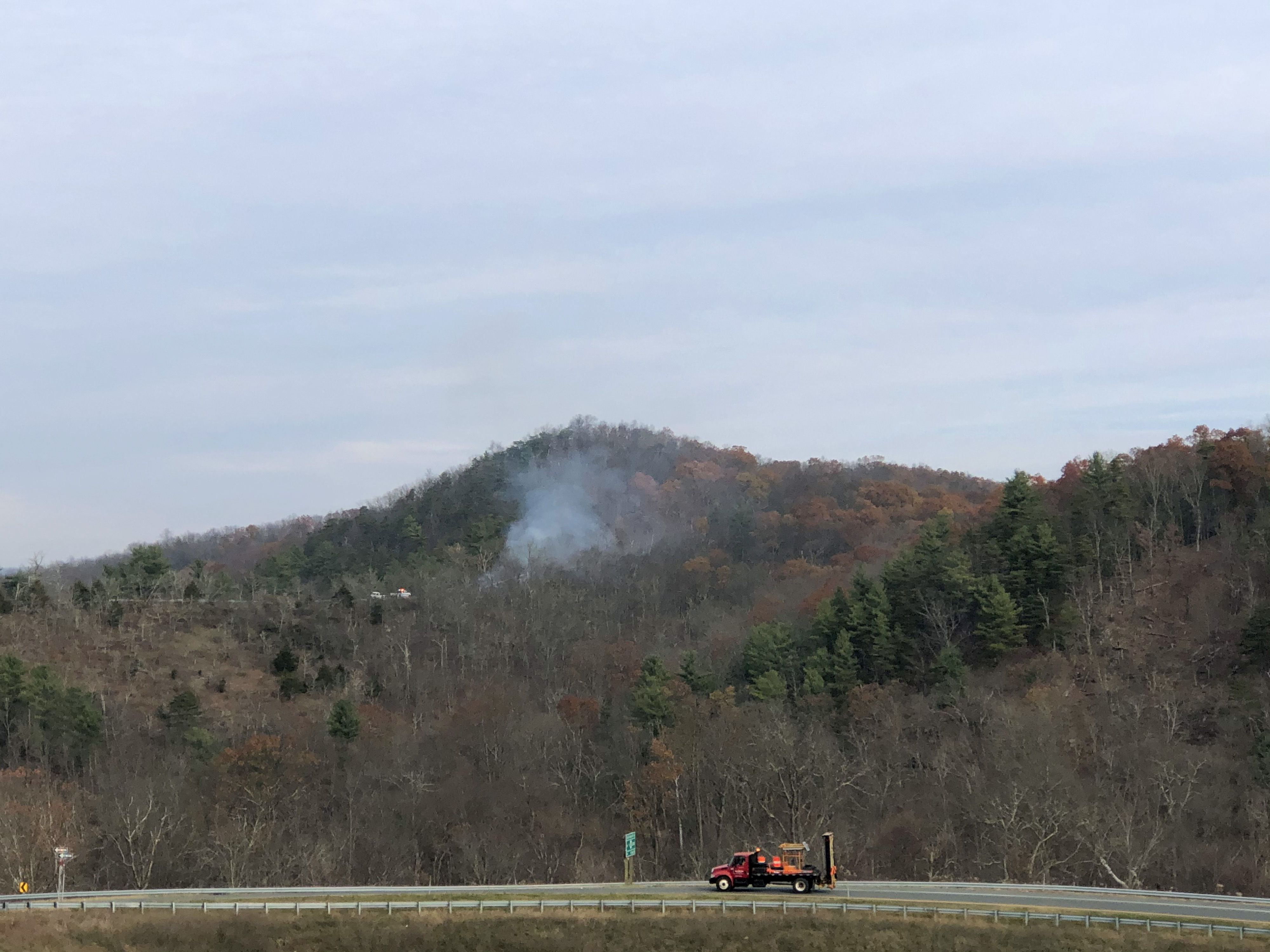 Smoke from a controlled burn rises above a green forested hill as cars drive by on the interstate highway that runs along the edge of the forest.