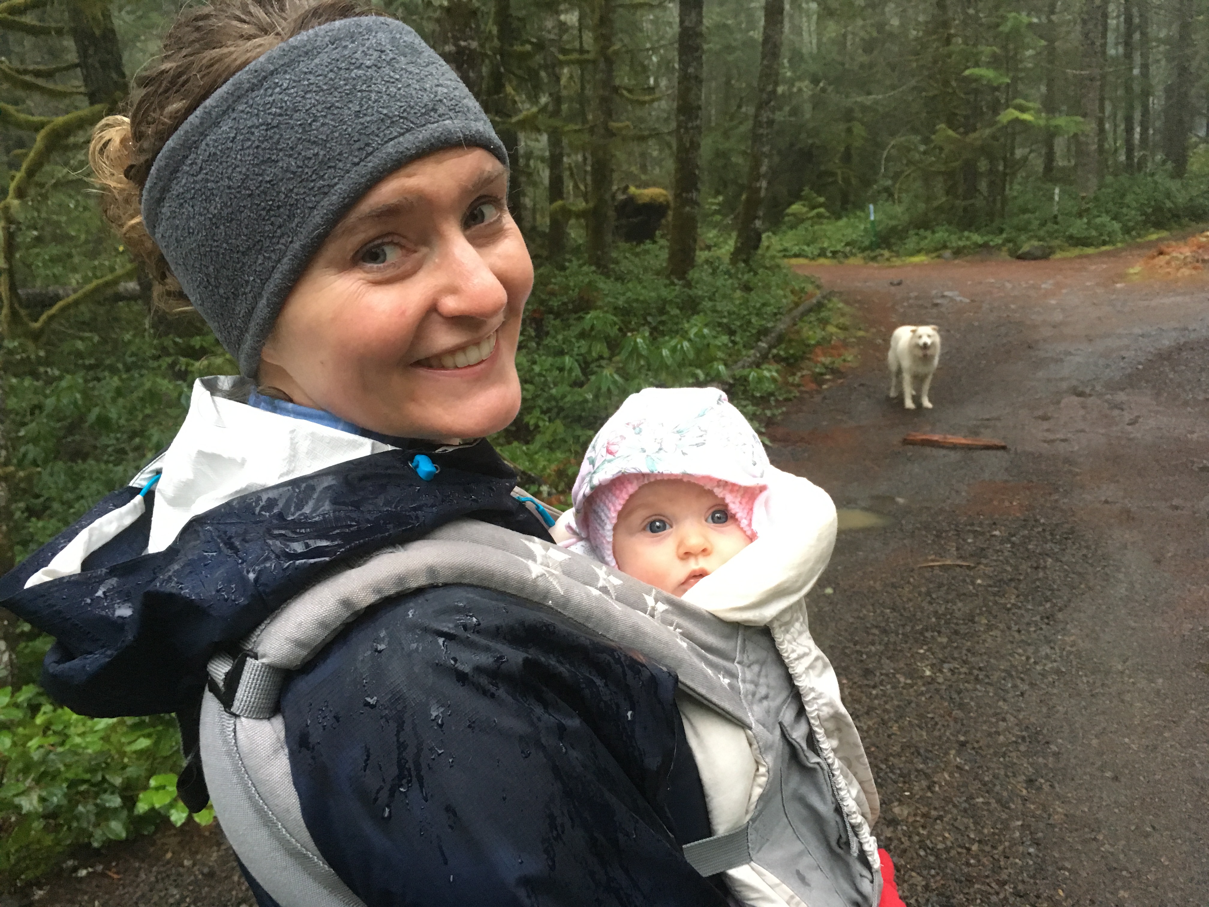 A woman holds a baby and smiles at the camera as they walk through the woods; a white dog stands on a dirt trail in the background.