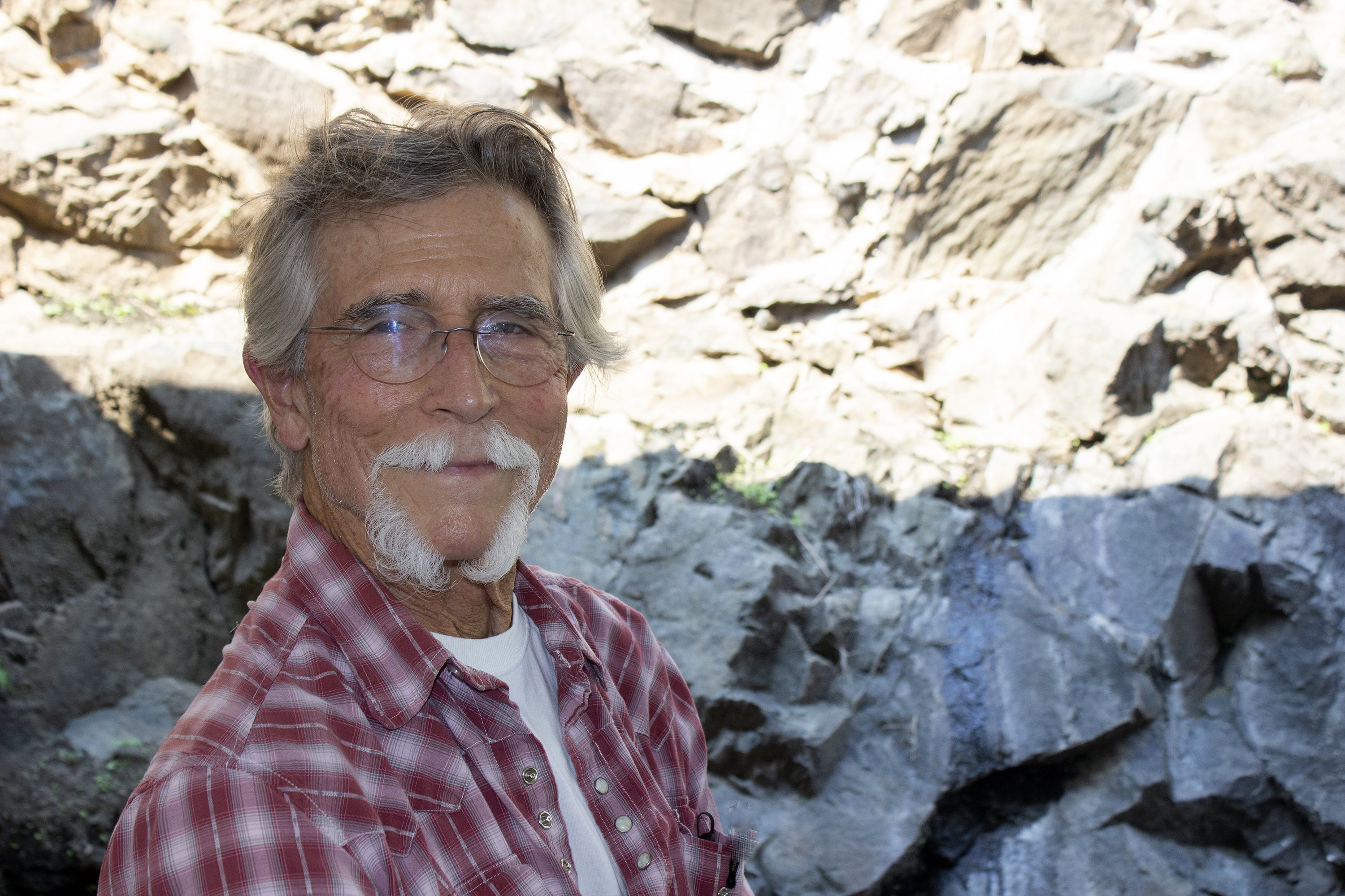 Portrait of a older man with graying hair and white beard. He is standing in front of a tall rock face.