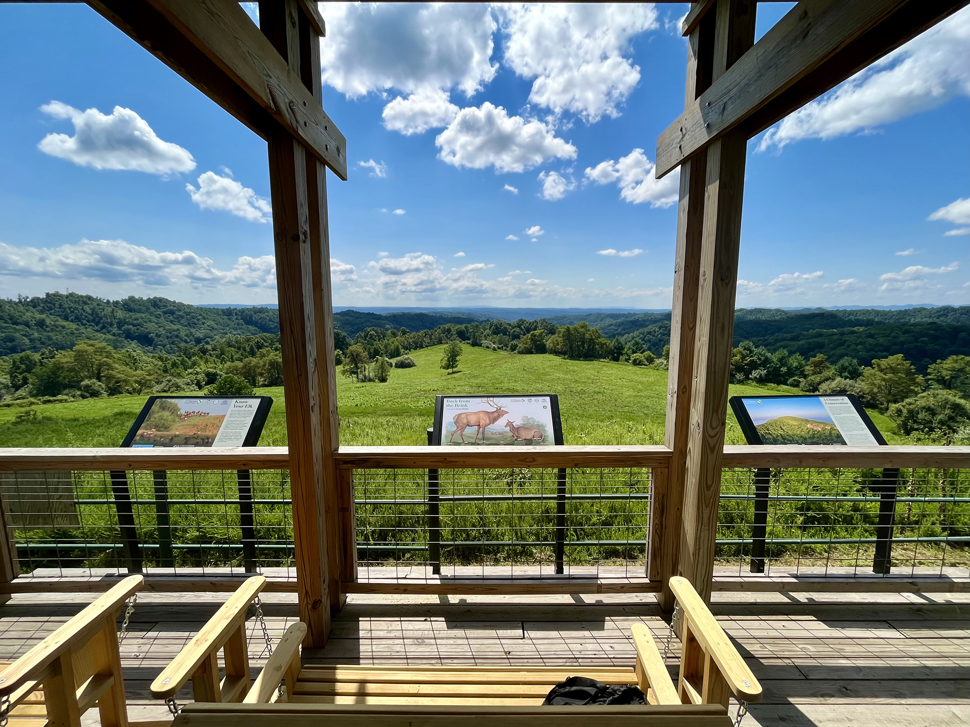 View from an overlook looking out on former minelands that have been converted to elk habitat. White fluffy clouds float over an open, green meadow. Three interpretive signs provide information.