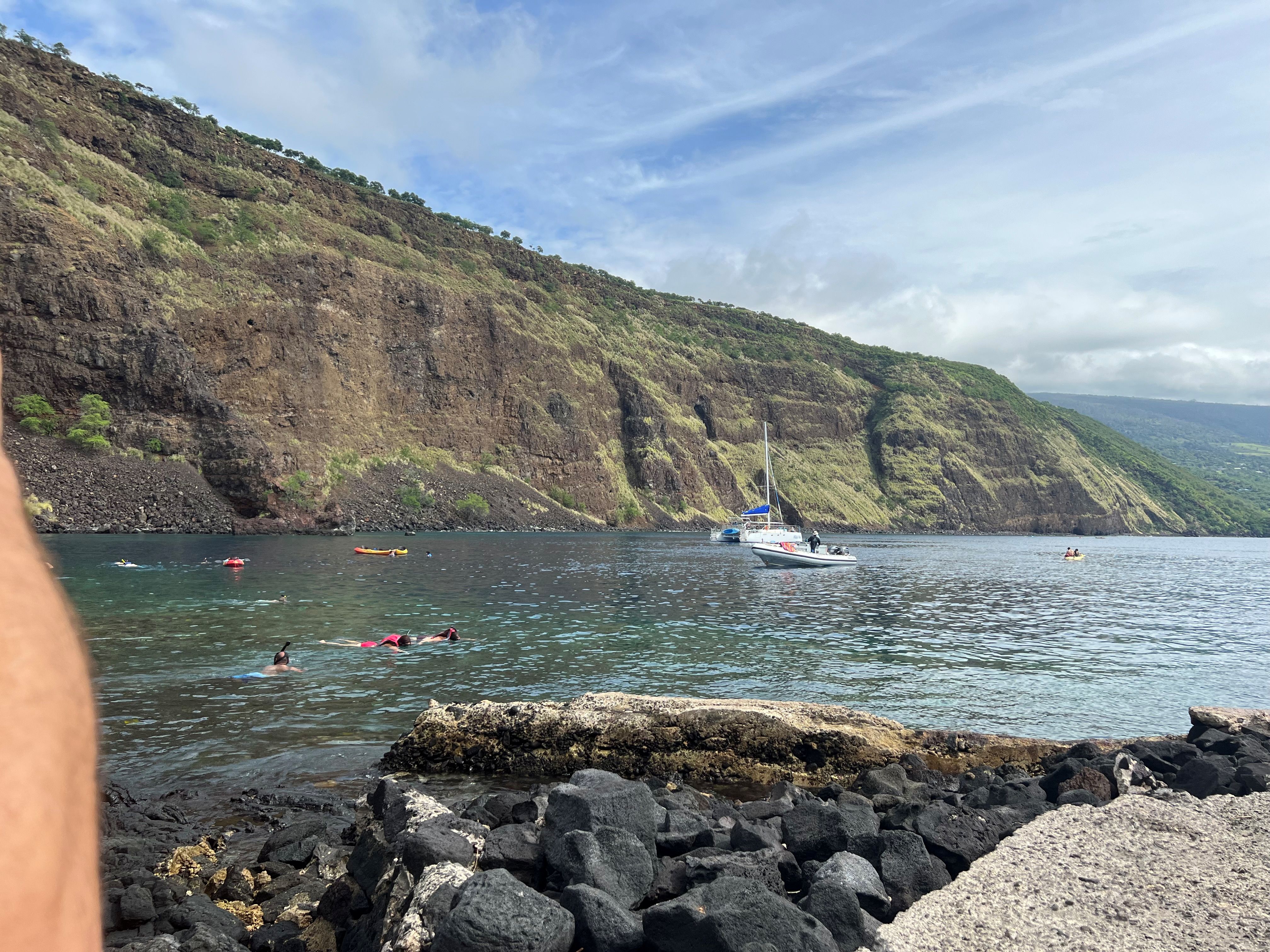 Boats, kayakers and snorkelers float in calm waters of small cove between cliffs and rocky shore.