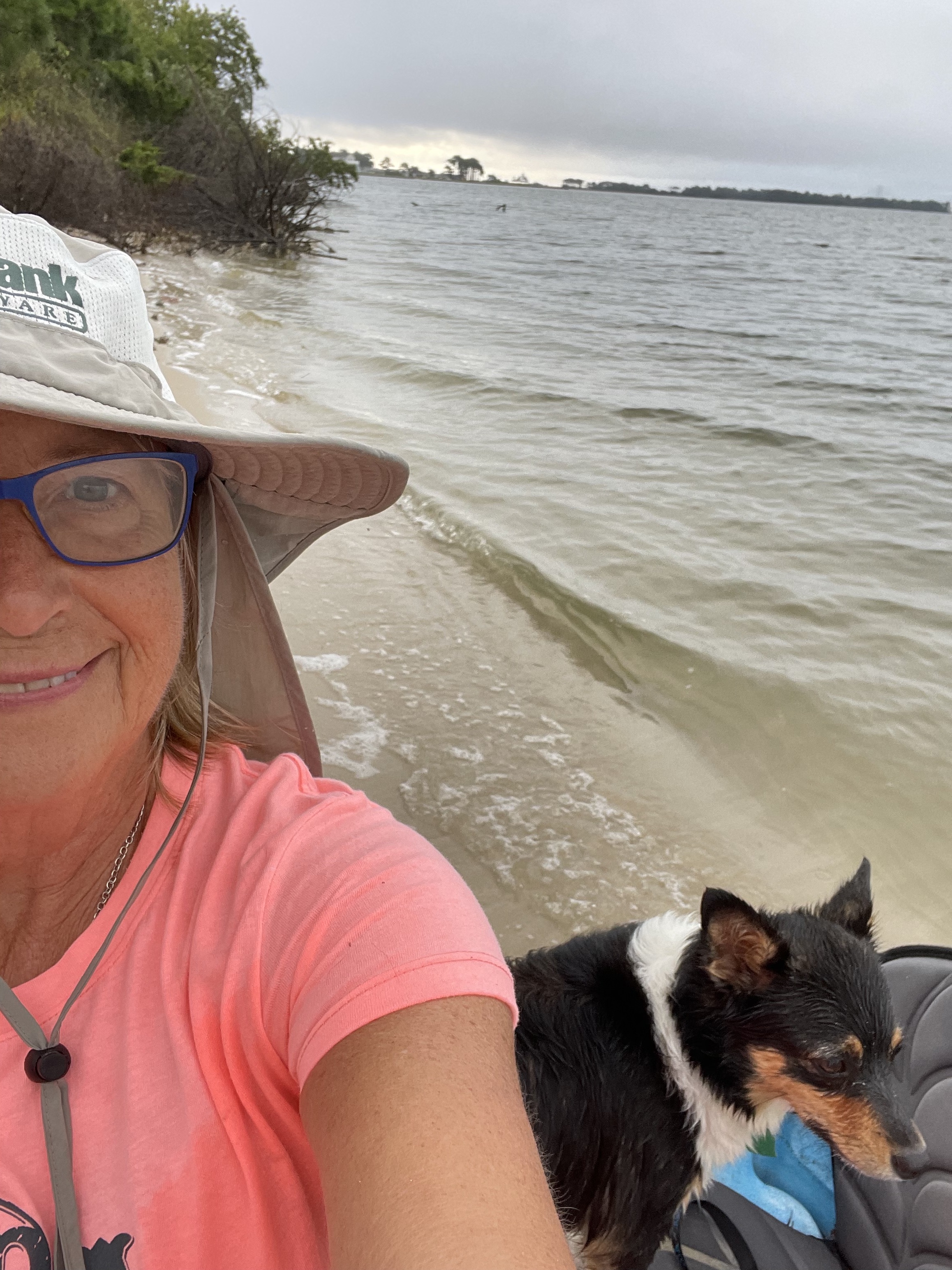 A selfie-style photo of Anita Janney smiling at the camera with a dog in front of a beach.