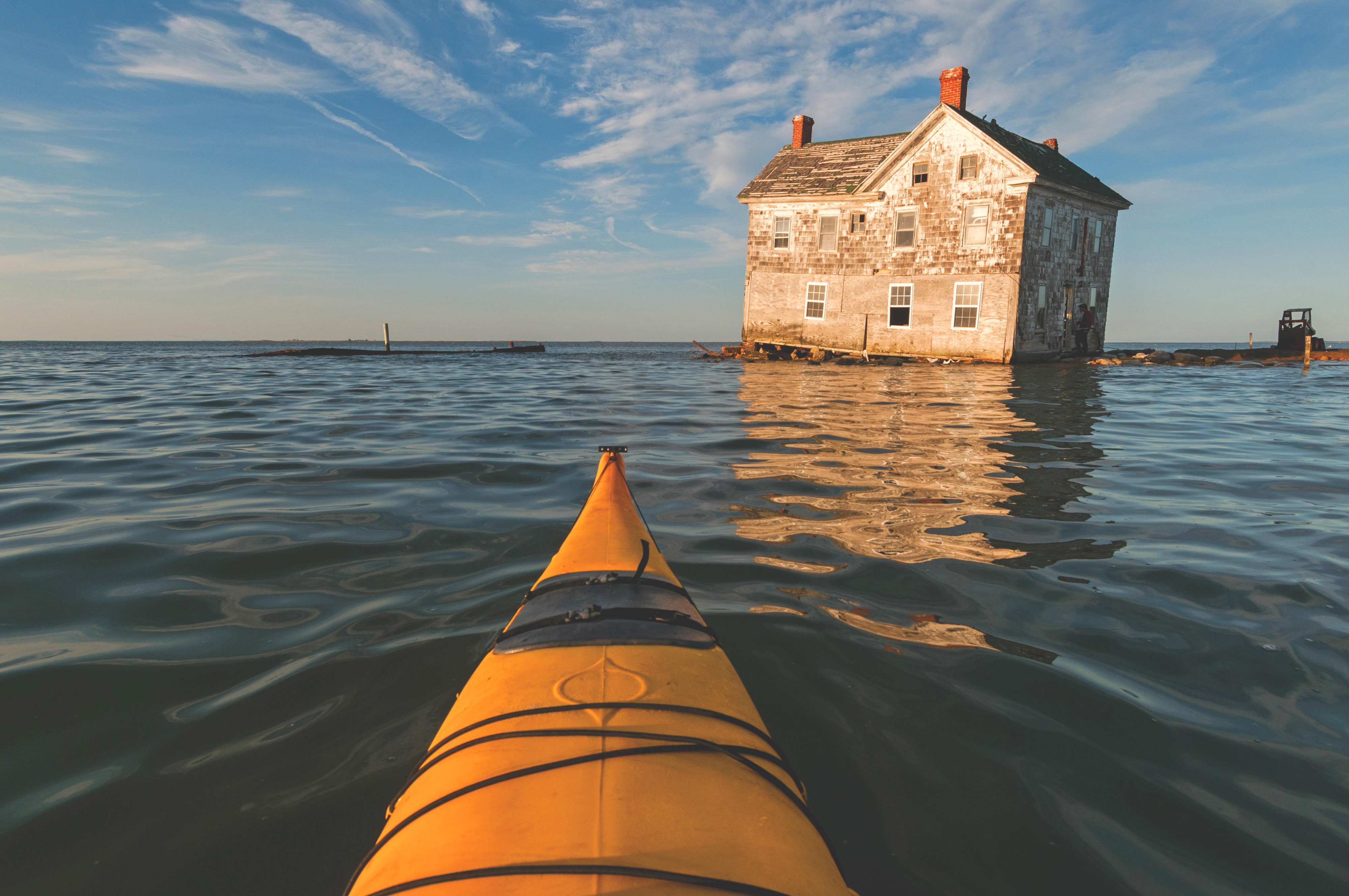 An abandoned farm house sits precariously at the edge of the water as the Chesapeake Bay slowly consumes it. The yellow nose of a kayak is visible in the foreground.