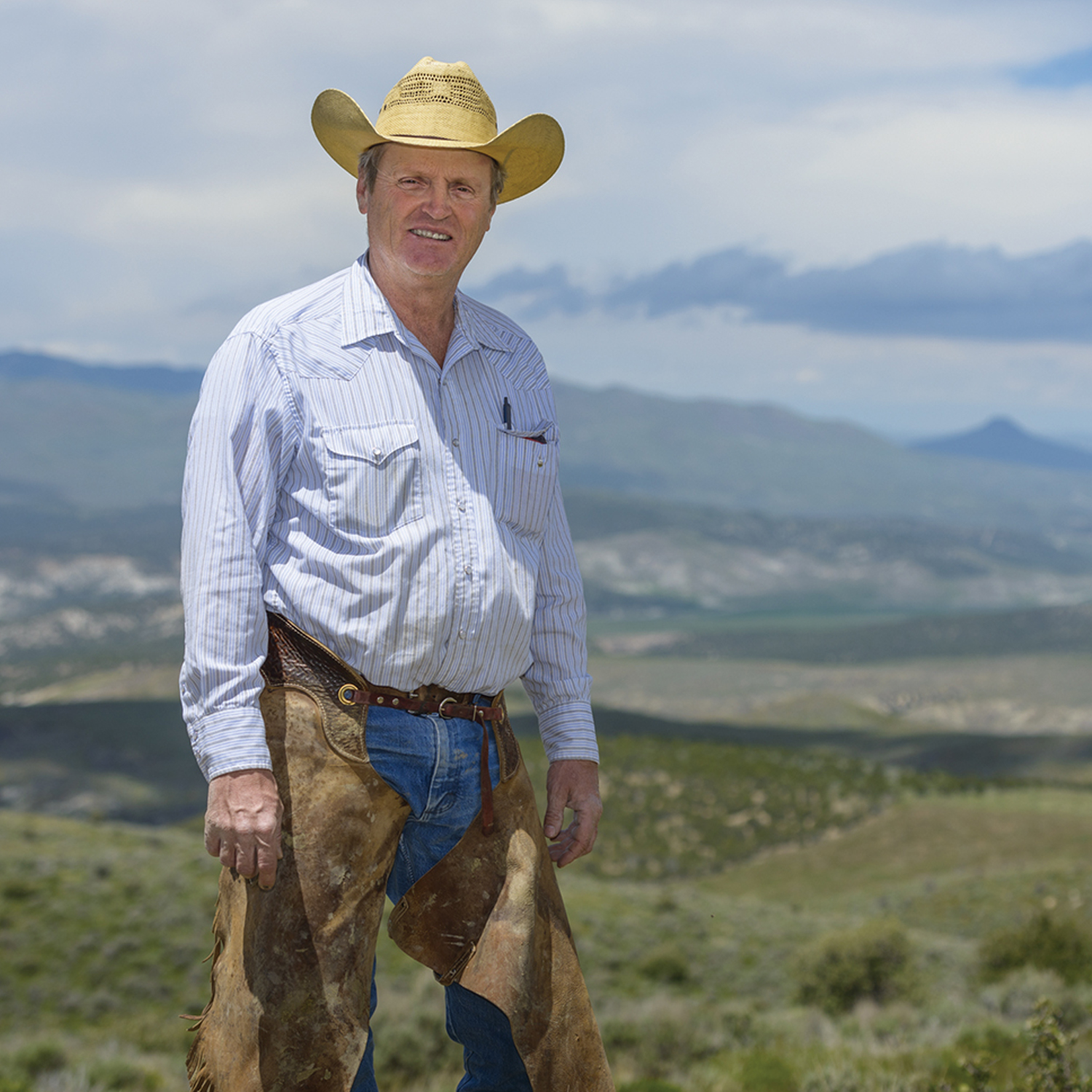 Jay Tanner wears a cowboy hat, blue shirt and brown chaps and looks at the camera. 