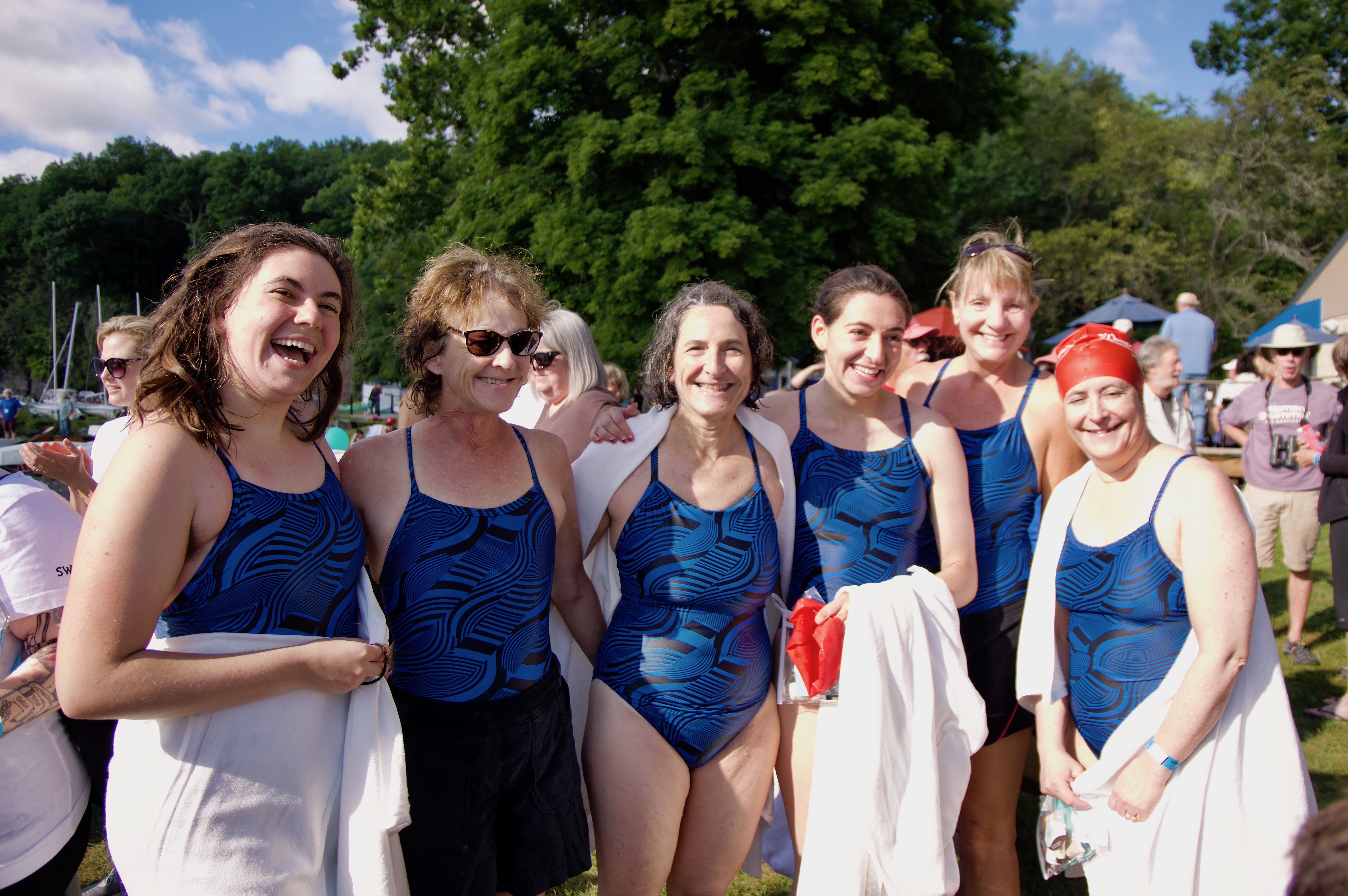 Six women wearing matching blue patterned one-piece swimsuits and holding towels and clothing smiling at the camera. A crowd of people are in the background. 