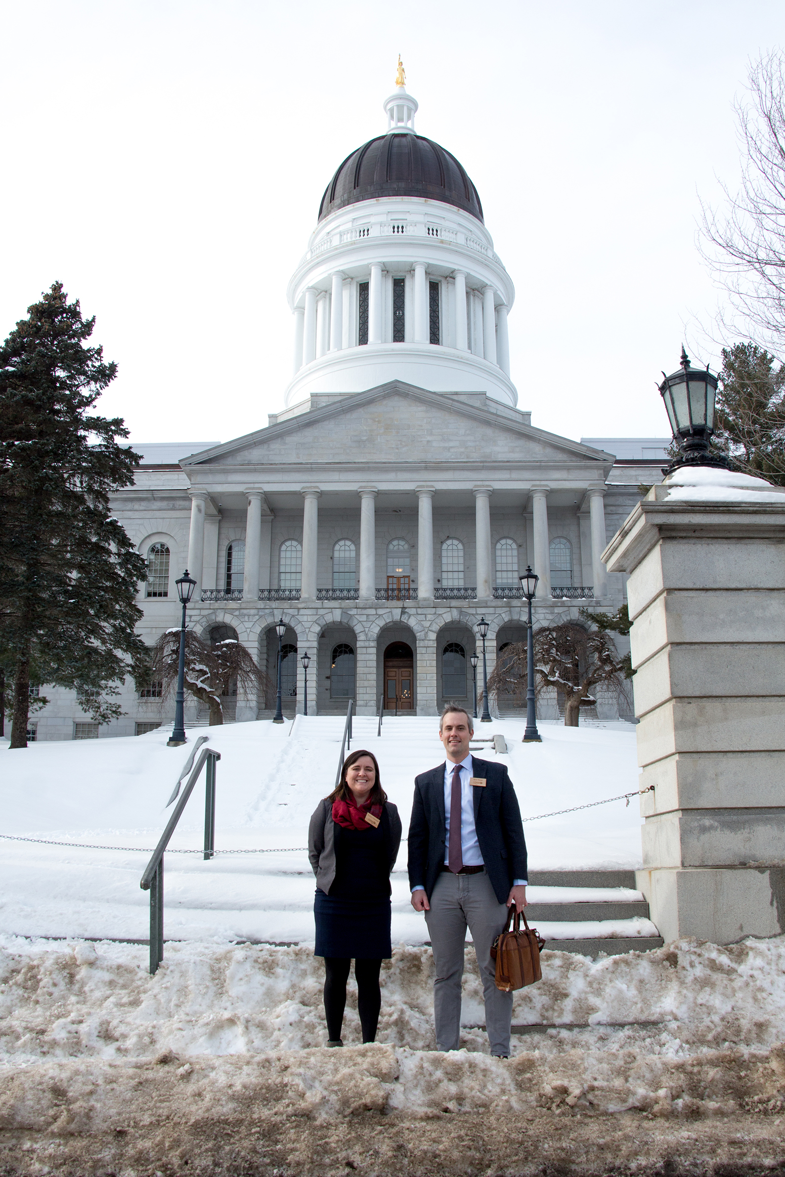 Kaitlyn Nuzzo and Rob Wood stand in front of the Maine State House on a snowy day.