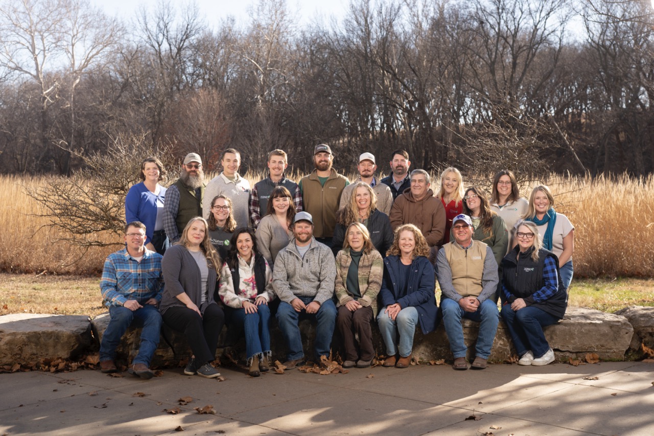 Group photo of about 20 staff members sitting and standing outside with forest in the background.