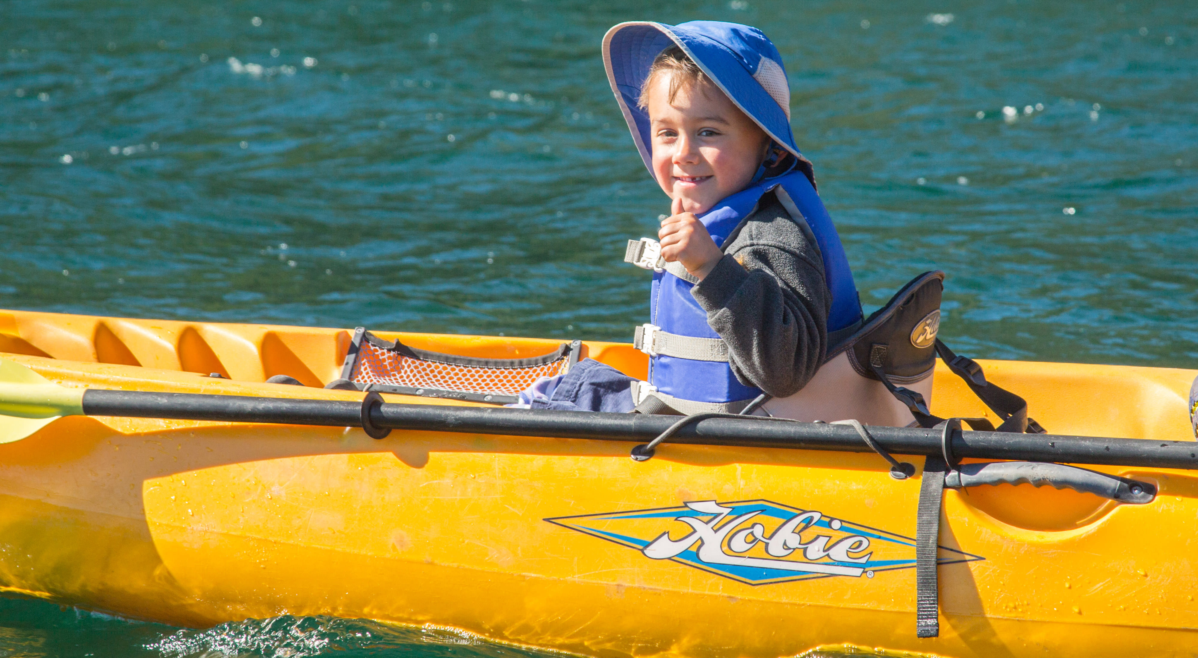 A young child sits in a yellow kayak on a body of water and smiles and gives a thumbs-up to the camera.