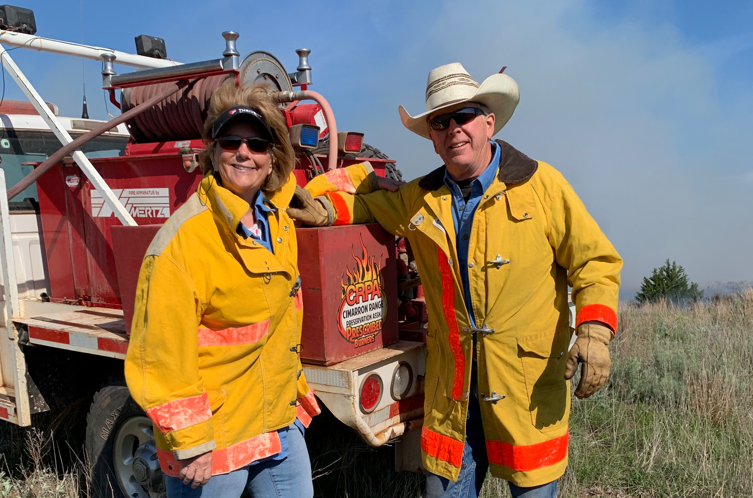 Man and women dressed in protective fire gear stand in front of a utility truck during a prescribed burn on a ranch.