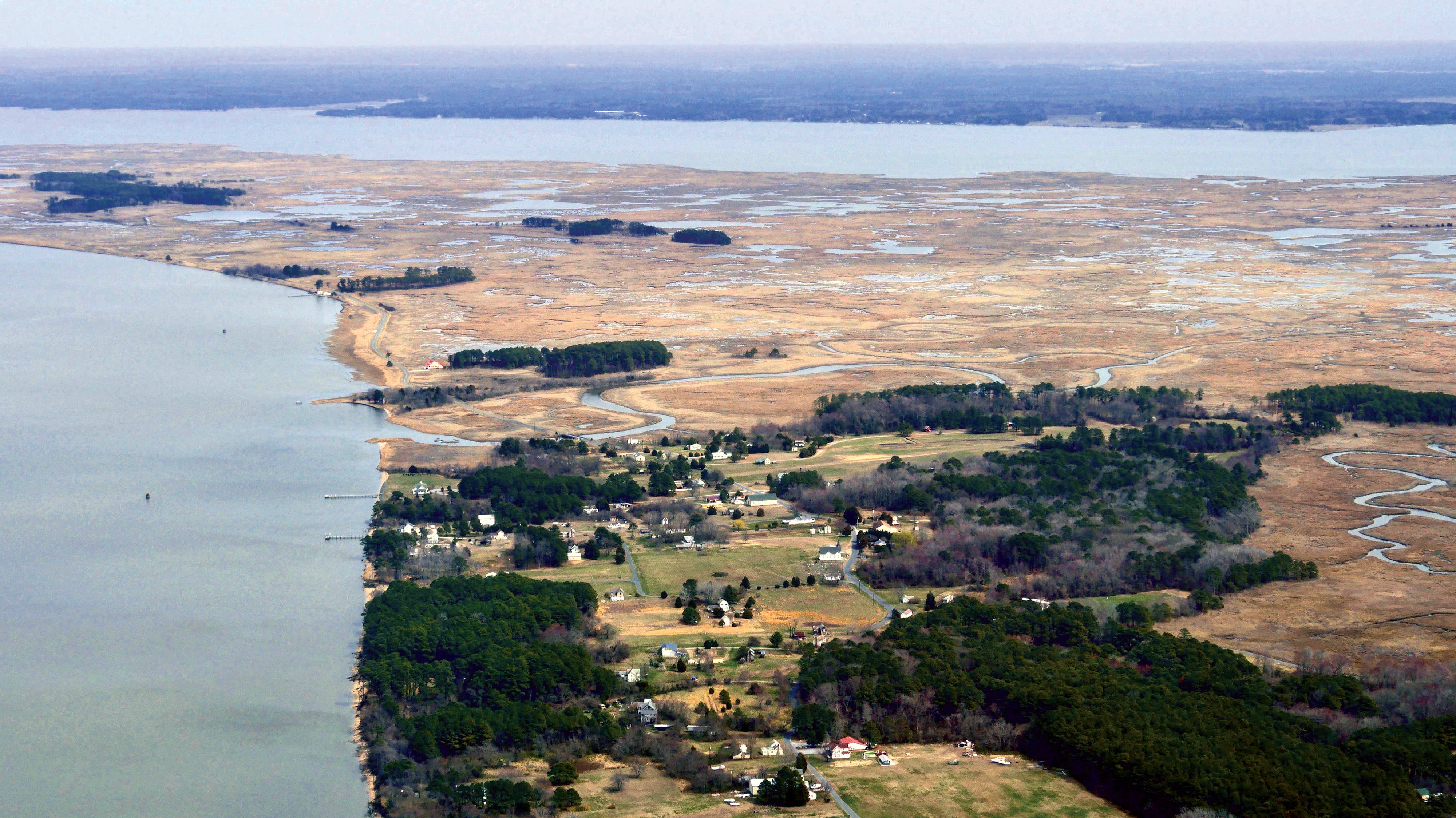 Aerial view looking down on a cluster of houses in a small coastal community. Open water faces the homes with marsh extending into the background.