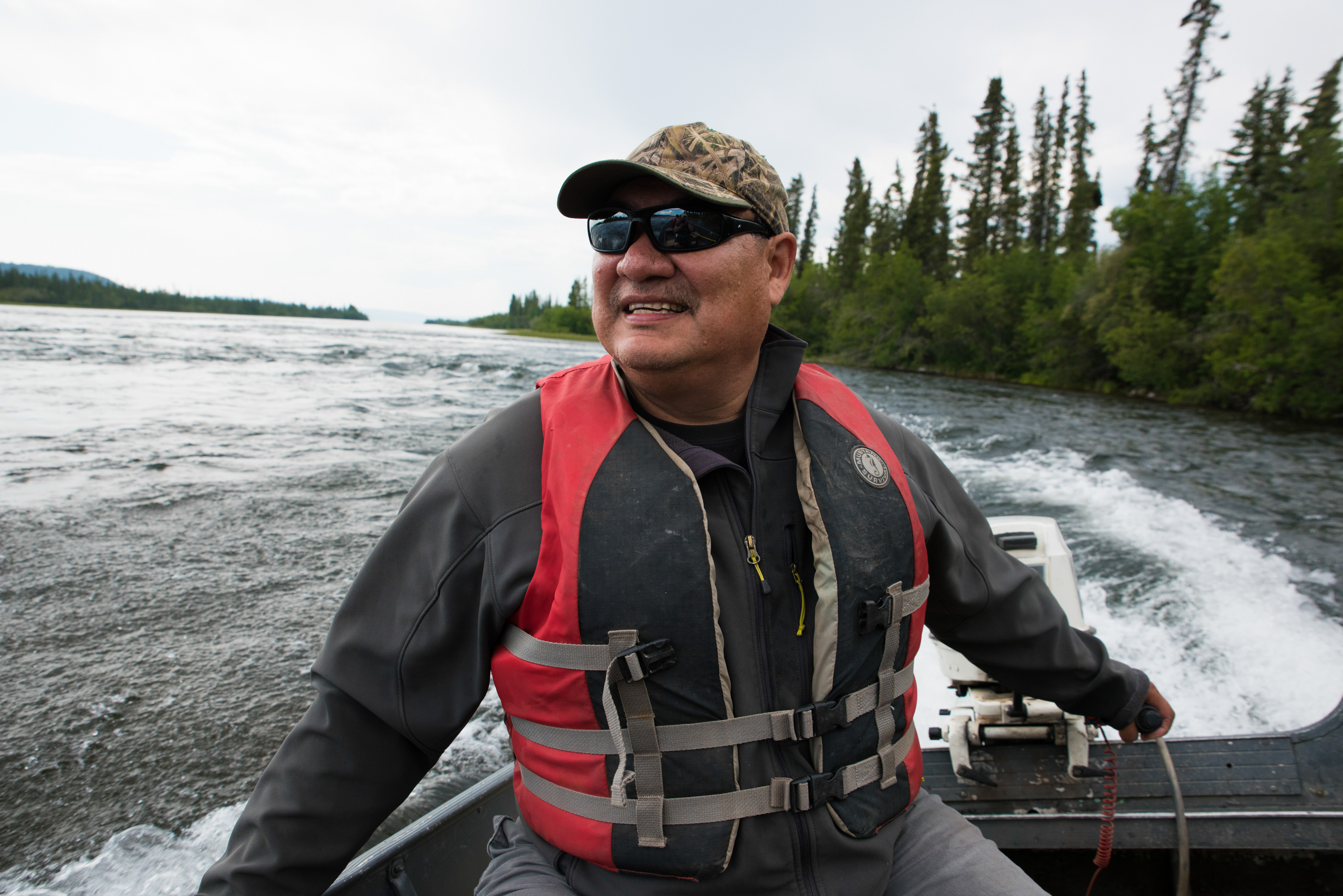 first nation man with sunglasses on a boat through a river surrounded by spruce forest in canada's boreal forest