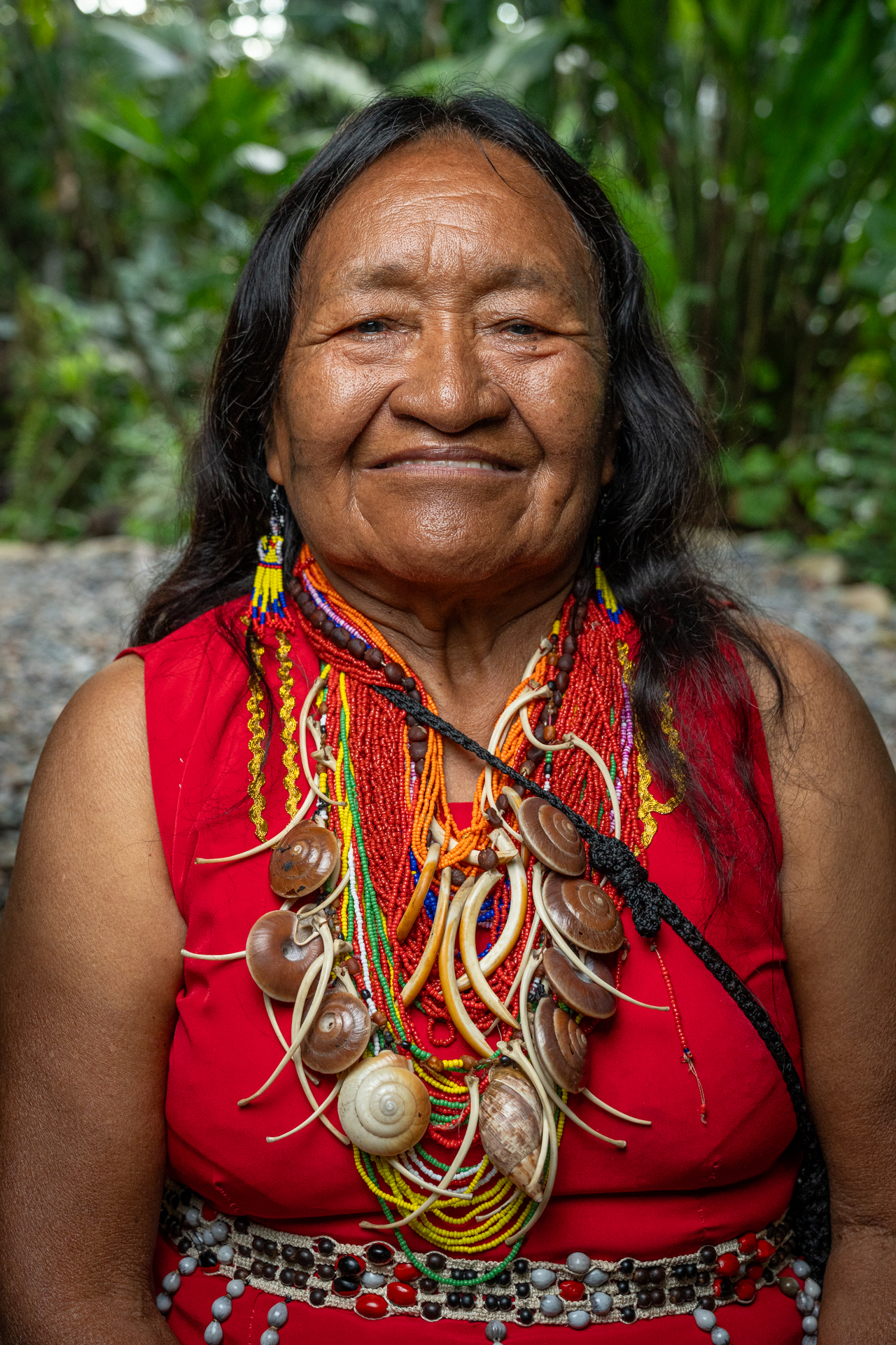 Retrato de mujer indígena madura, vestida de rojo, con collares de conchas, aros amarillos y el pelo oscuro y suelto. 