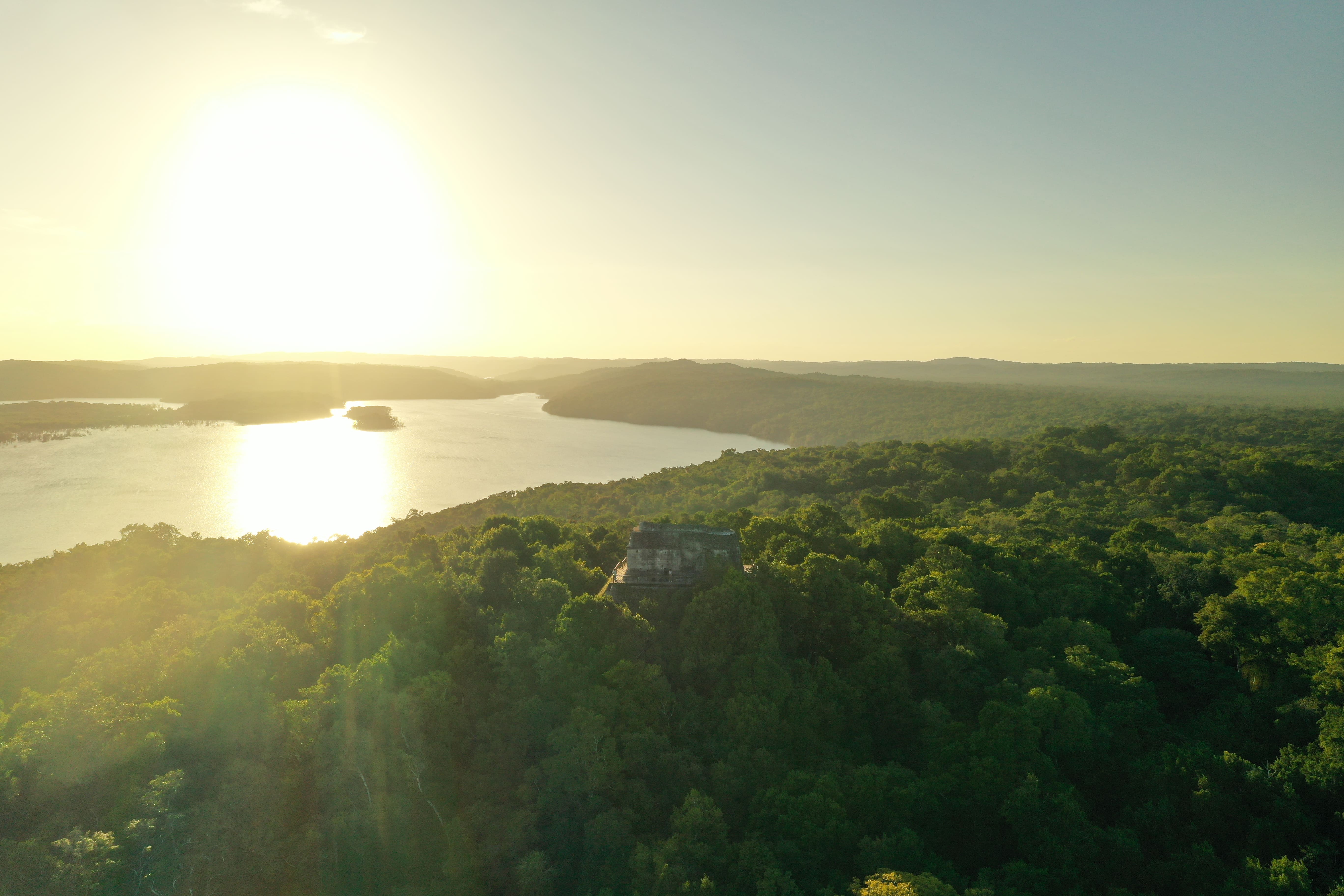 Vista panorámica en la Selva Maya al atardecer donde se observan pirámides entre los arburtos y un rio en el fondo.