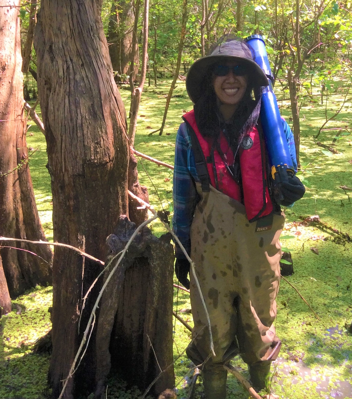 A woman wearing a floppy hat and sunglasses stands in the middle of a swamp.