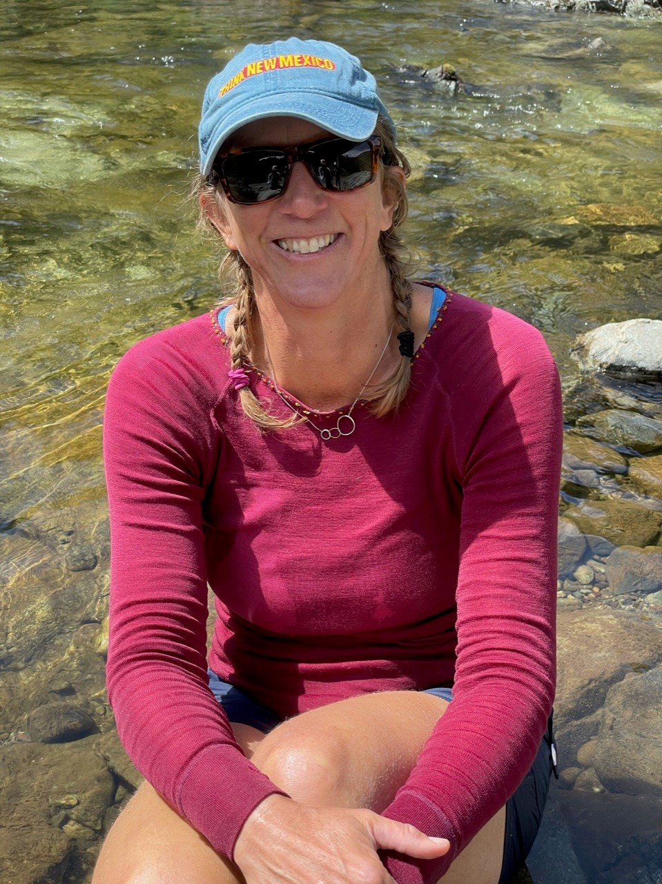 Woman with braids wearing a red shirt, blue ball cap and sunglasses.