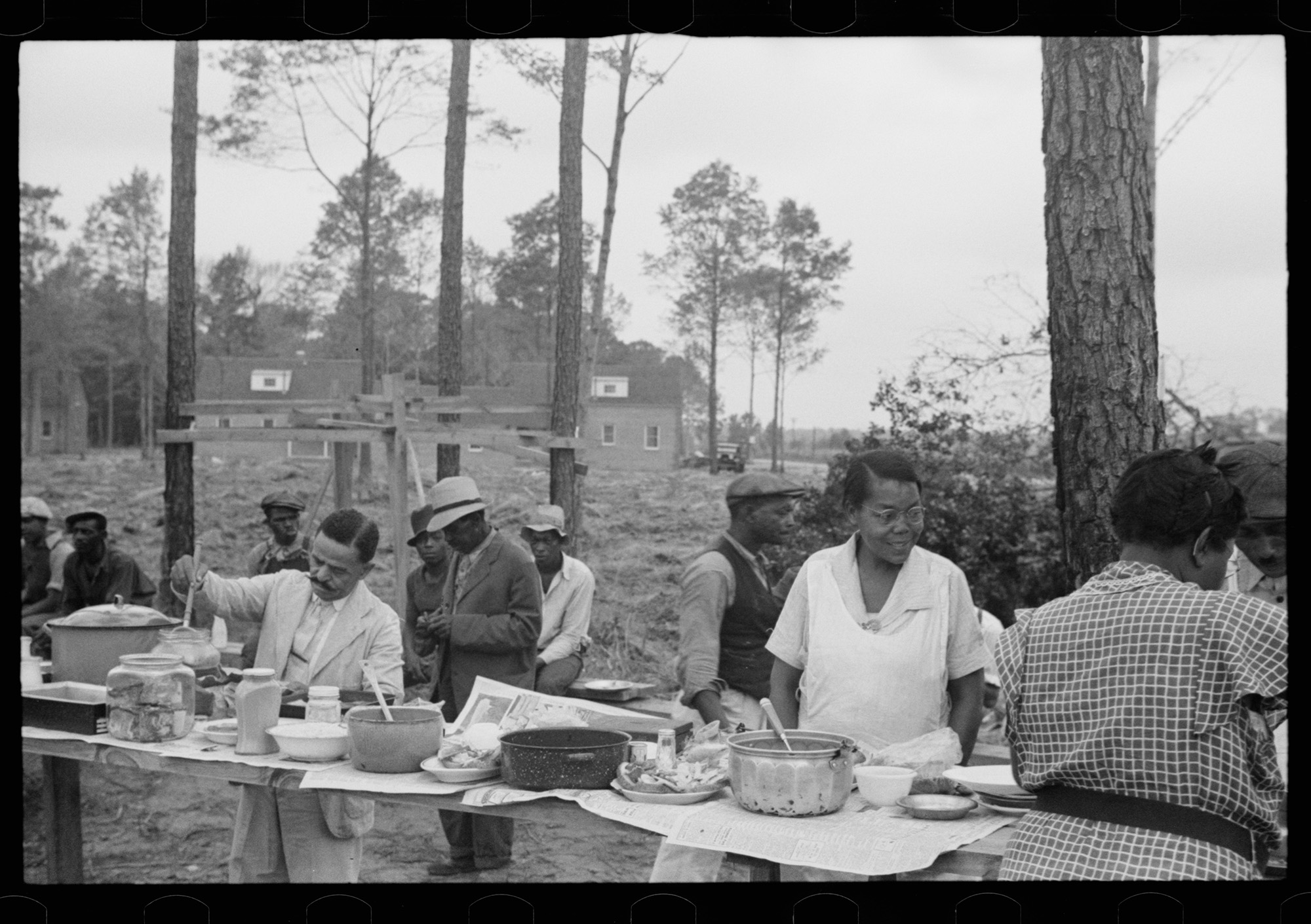 Black and white photo from the 1930s. A group of people enjoy an outdoor meal. Dishes are set up buffet style on rough cut boards. 