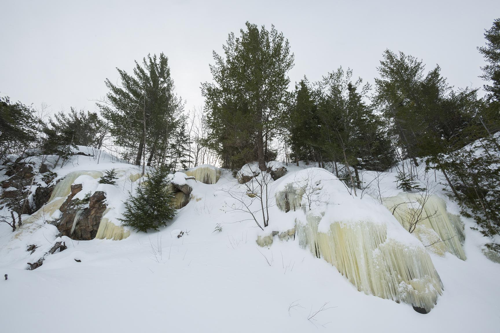 A bedrock outcropping is blanketed in snow and ice. Trees dot the landscape.