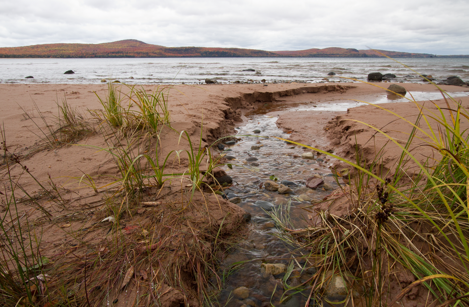 A sandy and rocky shore with a small inlet of water making its way to the lake.