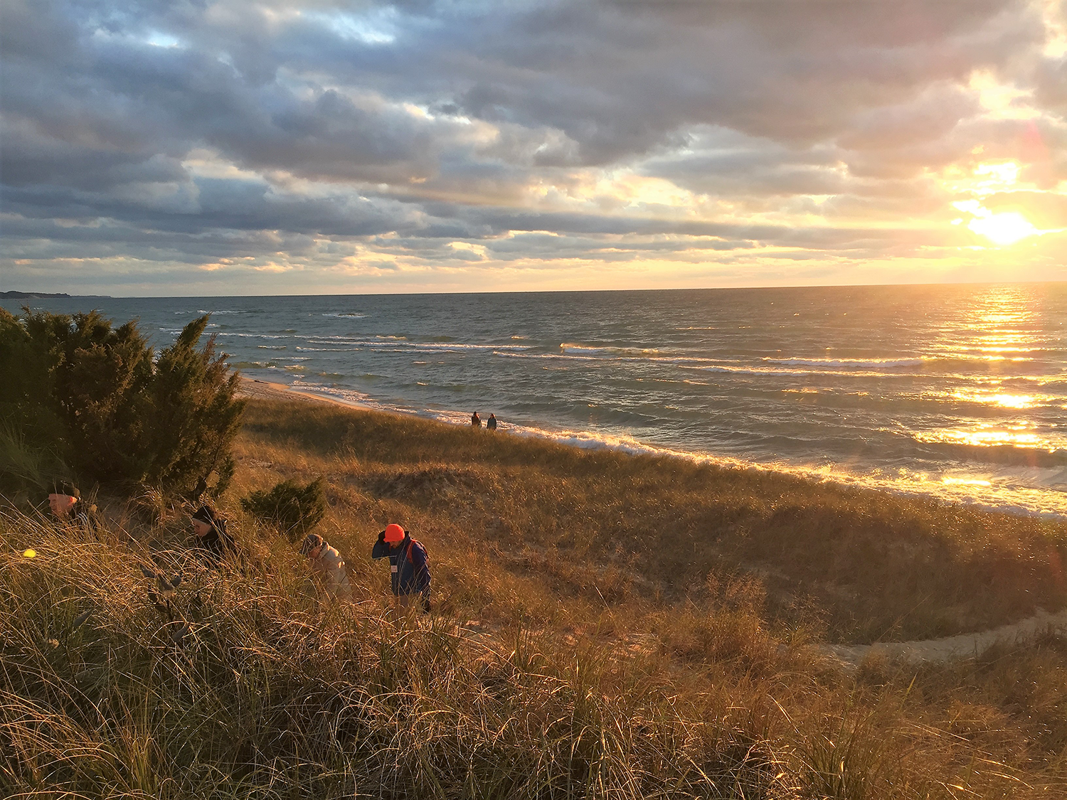 Hikers climb up a grassy incline as the sun sets over Lake Michigan.