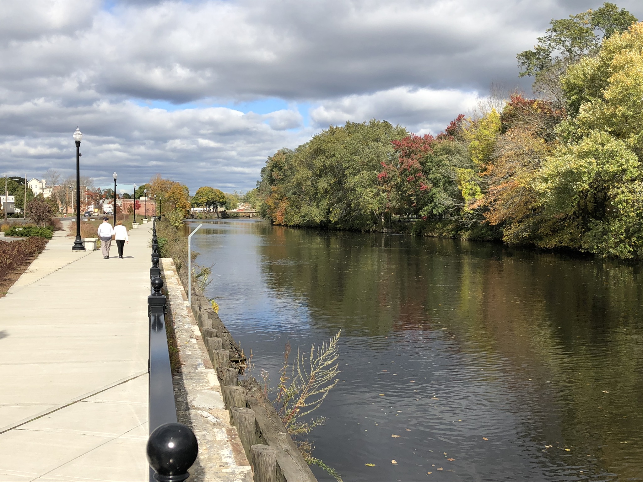 The Taunton River, with the riverside walkway on the left and trees on the right. A couple walks down the walkway in the distance.