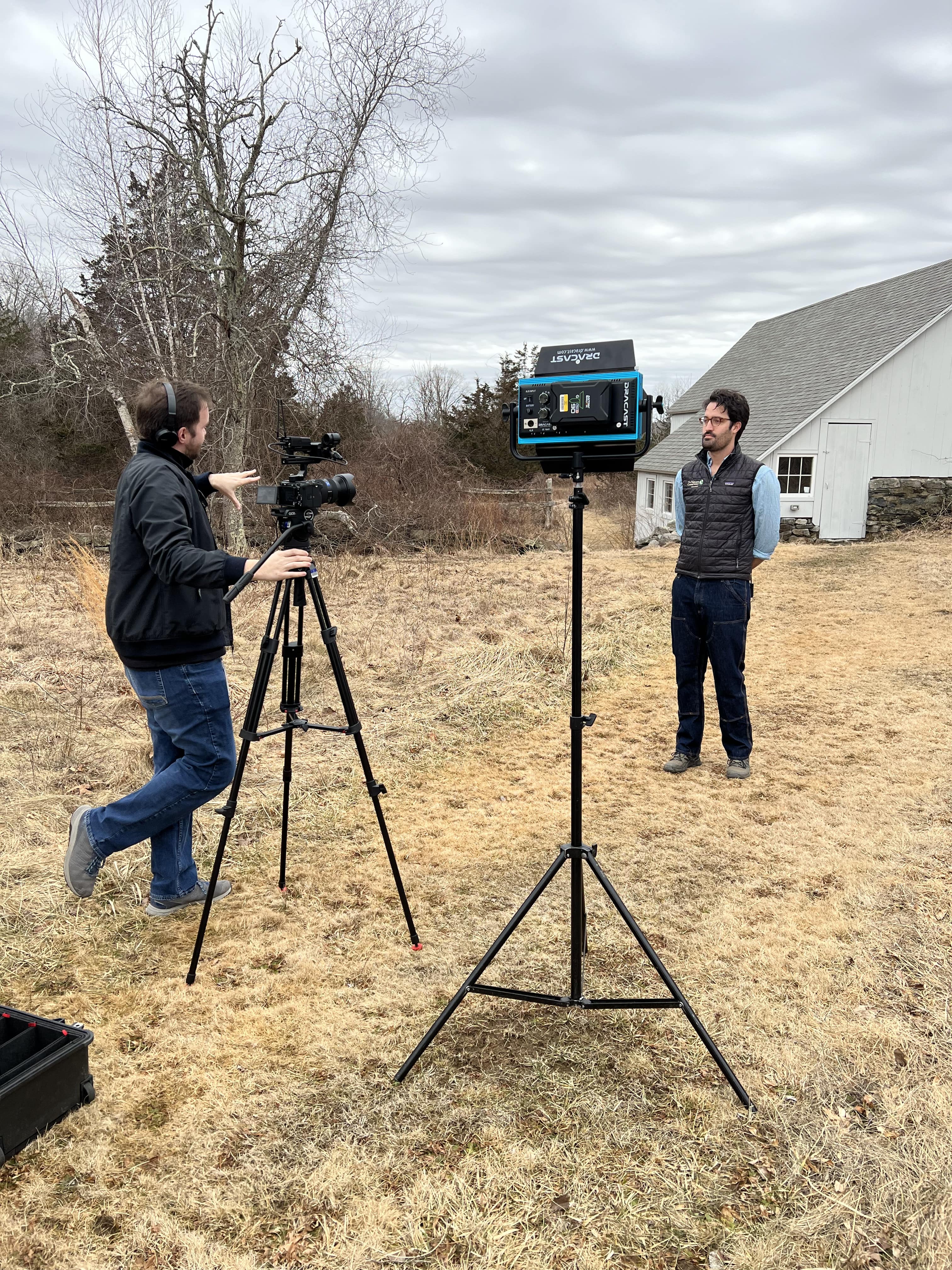 A man speaks to another man standing behind a video camera.
