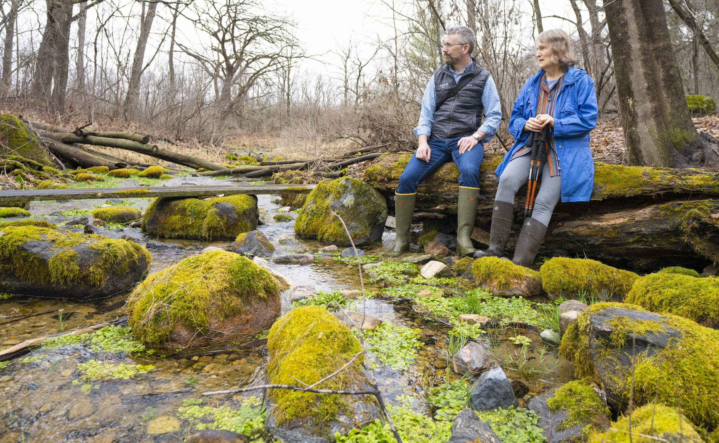 A man and woman in knee-high rubber boots sit on a log with their feet in a shallow, rocky stream.