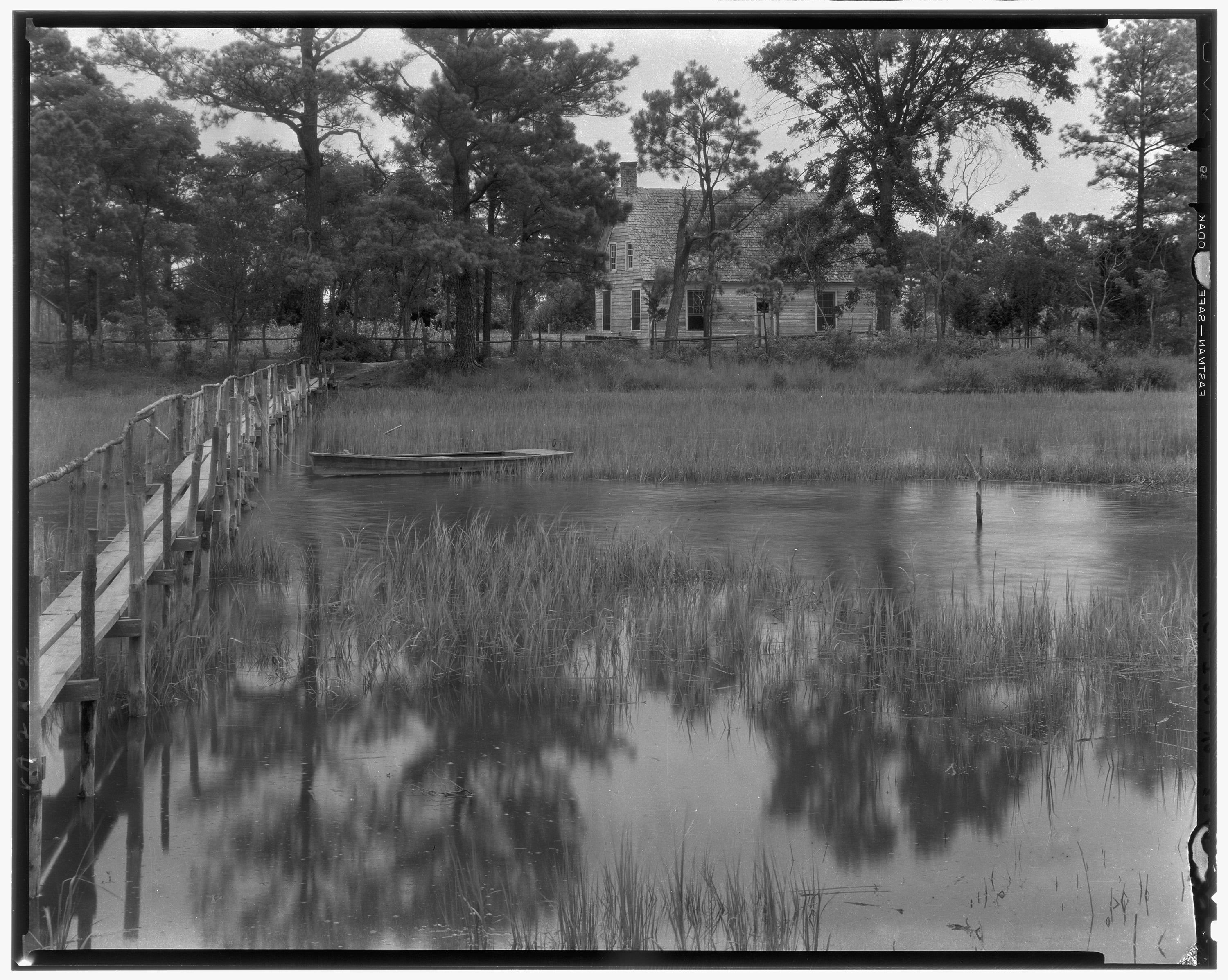 Vintage black and white image of an abandoned single story house nestled in the trees at the edge of a wide creek. 