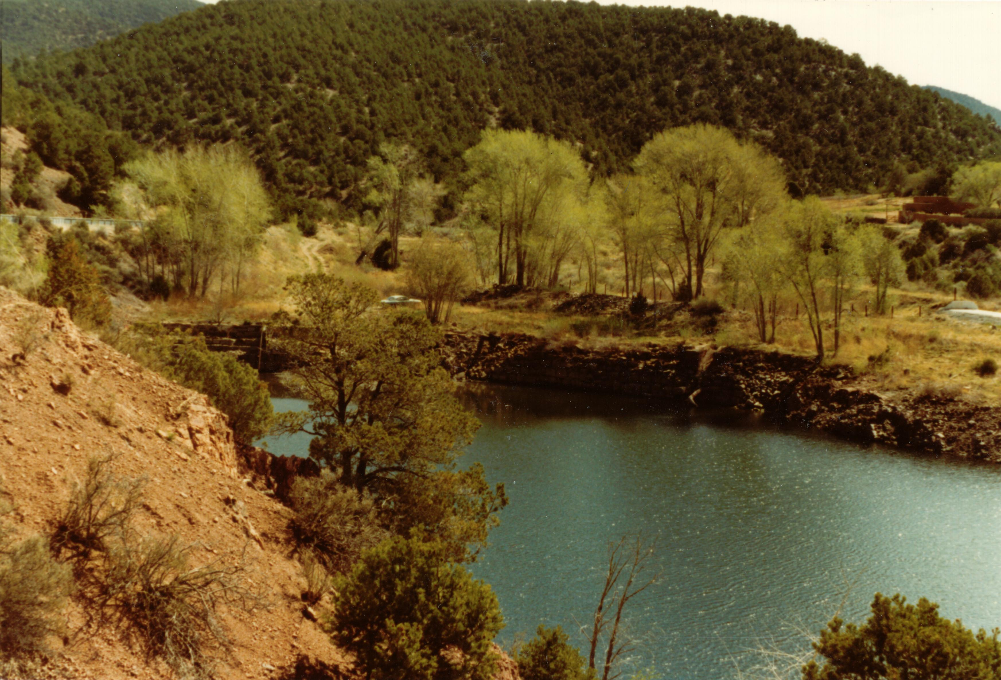 Old photo of stone dam on river in front of forested mountain.