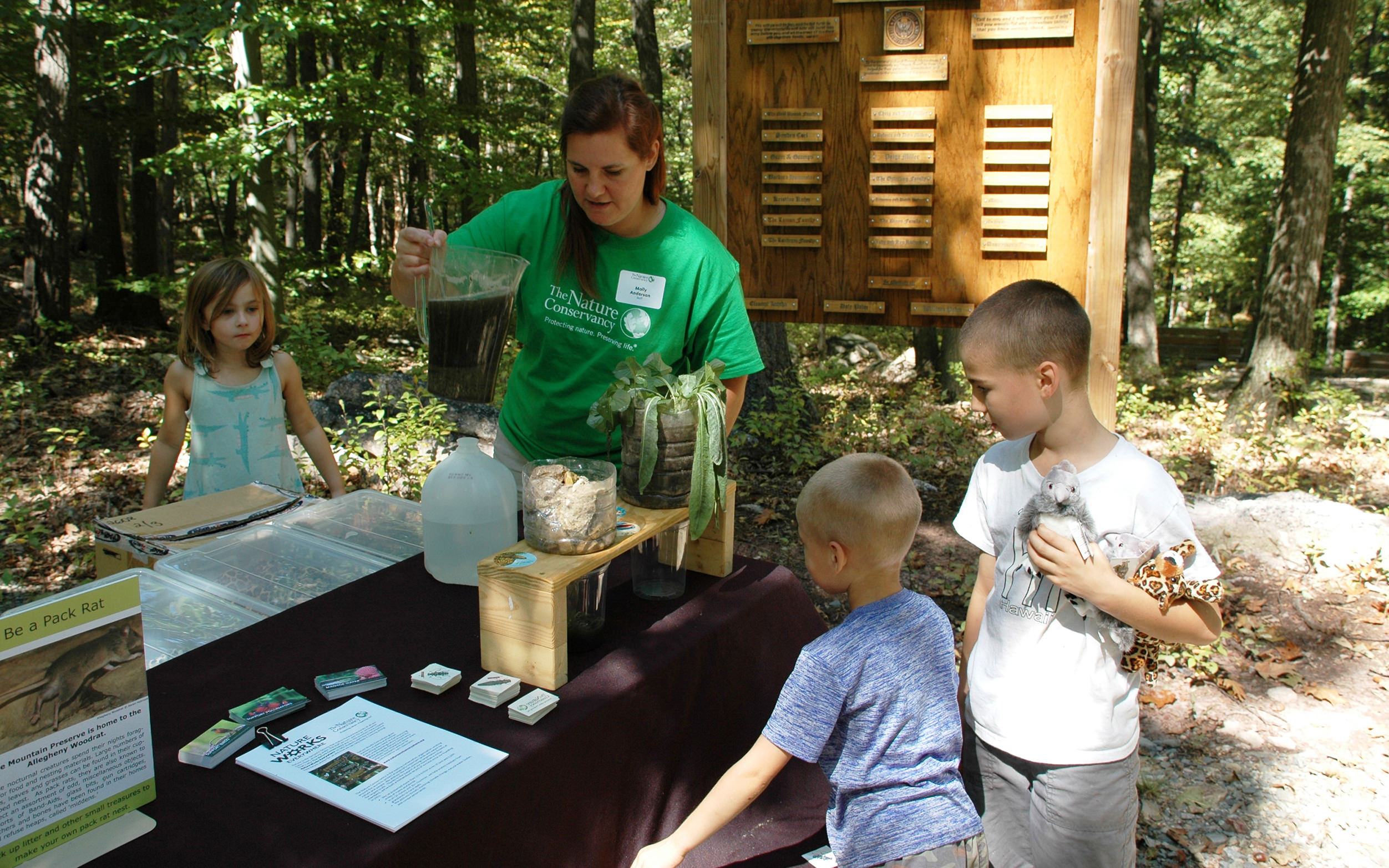 A woman stands at a table at an outdoor visitors center. She holds a pitcher full of dirty water showing three children a water quality testing experiment.