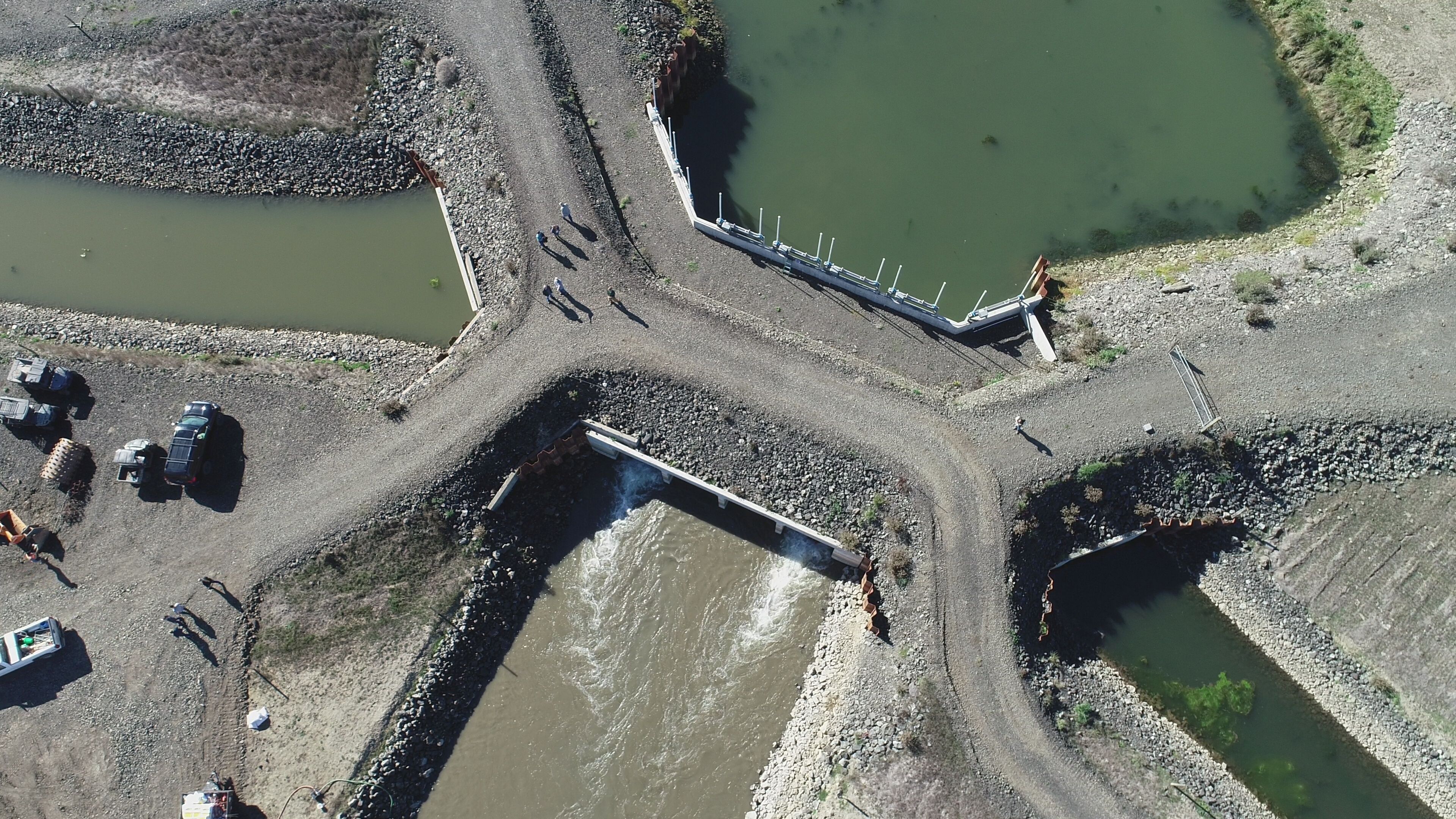 Aerial view looking straight down on gravel roads and parking areas, with several channels of water coming together, and water flowing through a white tide gate in the middle.