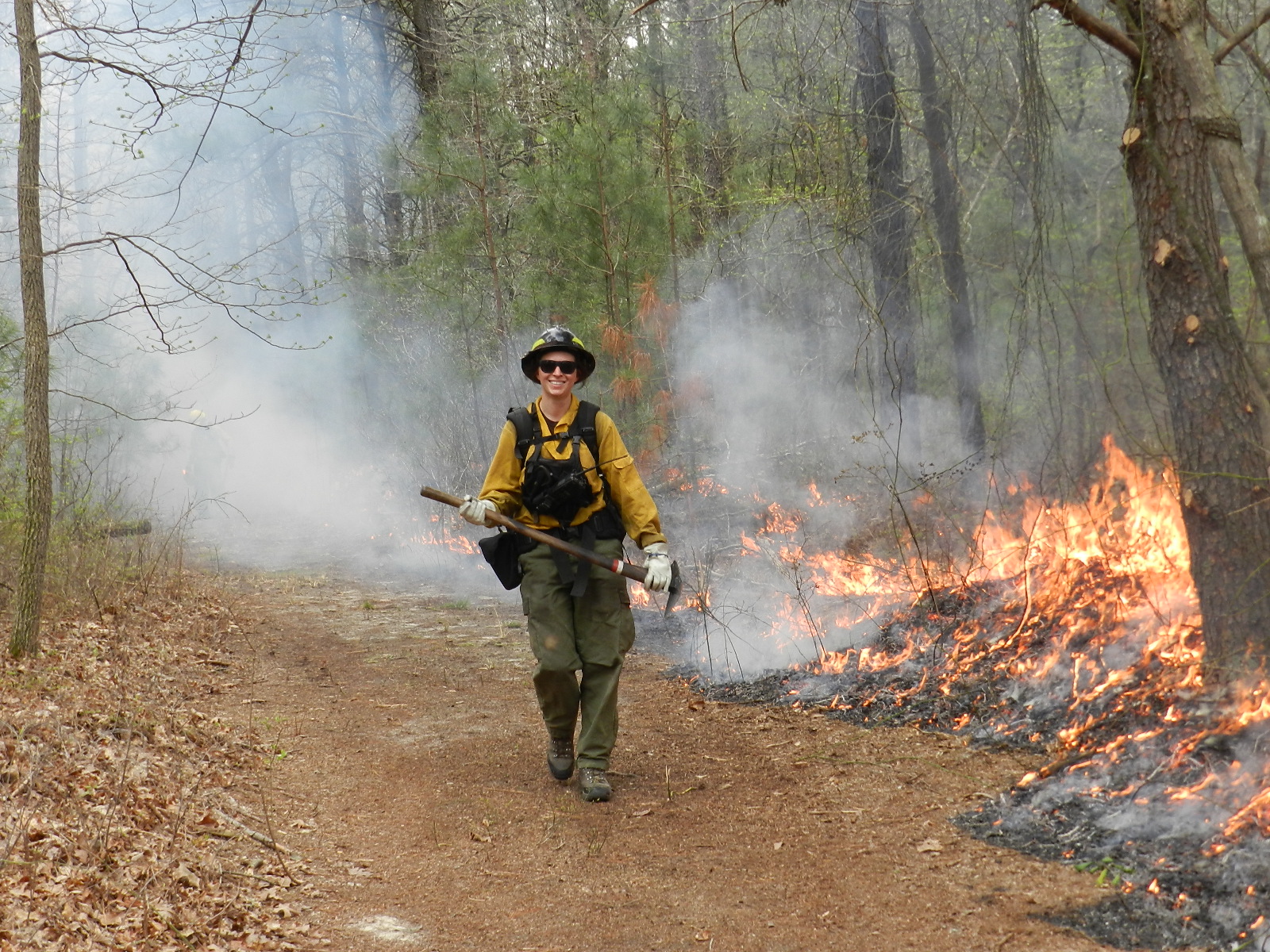 A person dressed in fire protective gear and holding an axe walks on a dirt path in a forest as a controlled fire burns behind them.