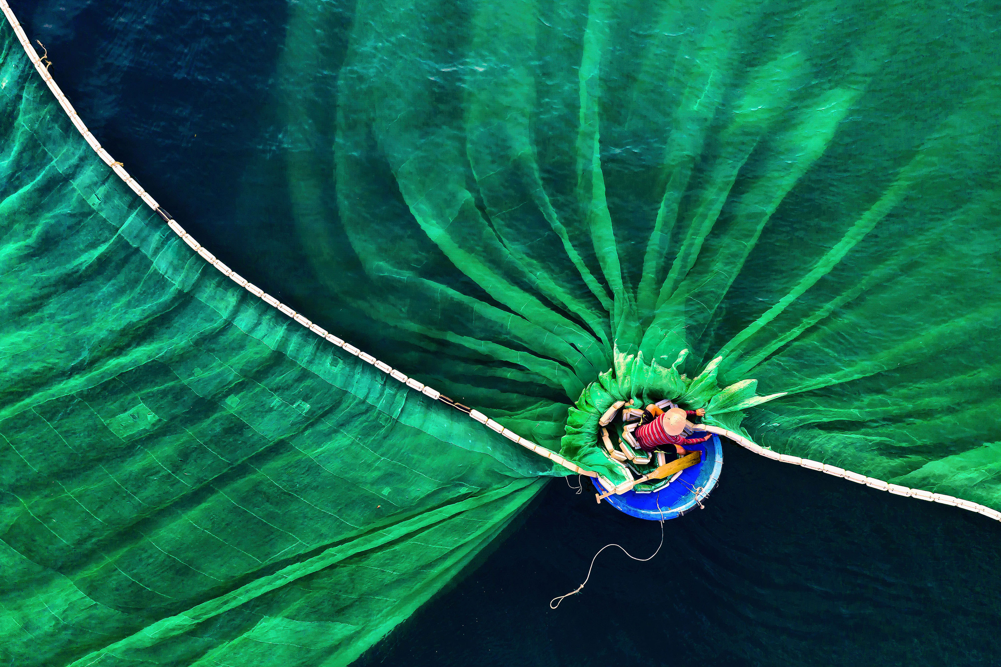 A person in a small circular boat casts a massive fishing net into the water.