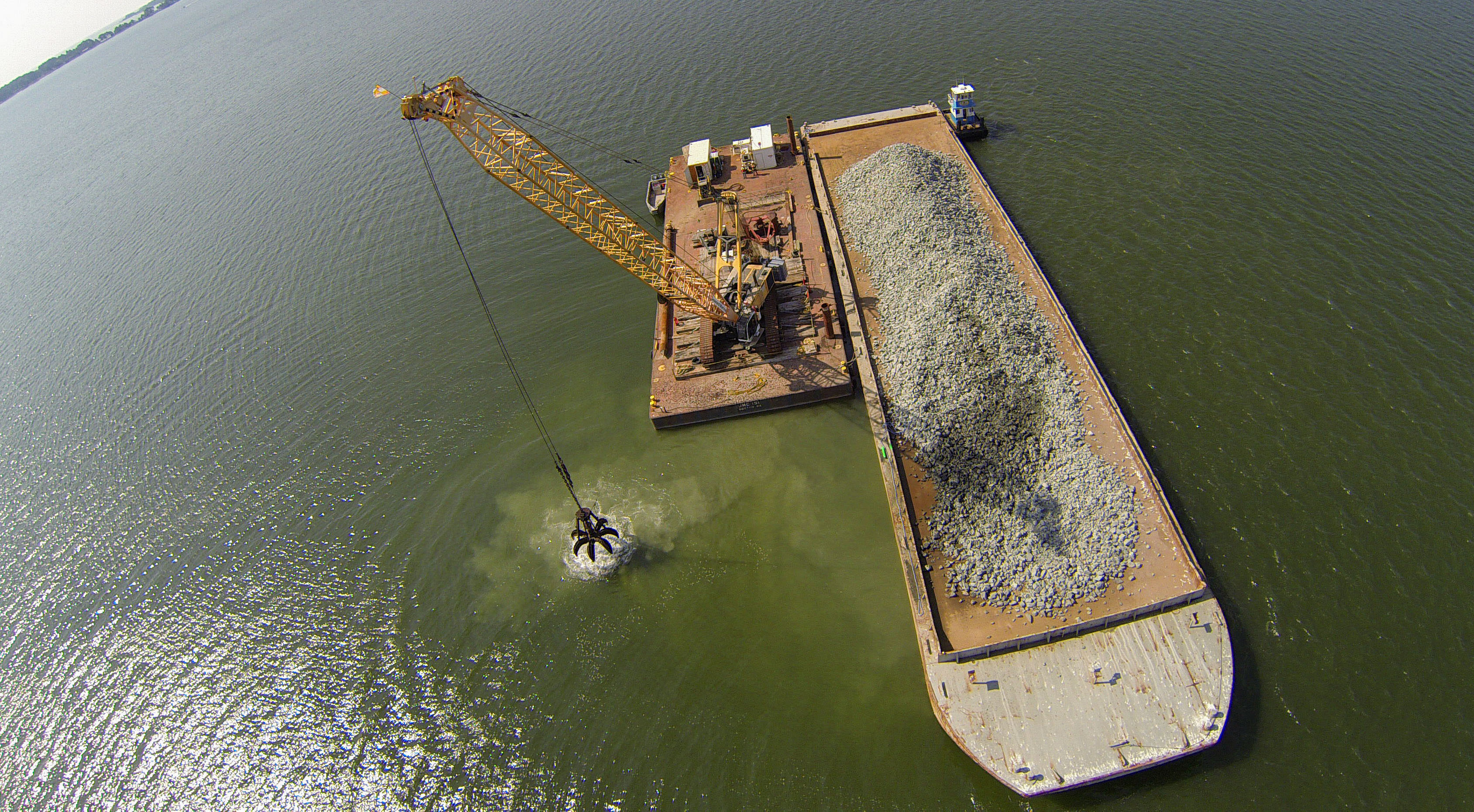 Aerial view looking down on a floating barge in the Piankatank River. A large crane is being used to scoop up chunks of granite rock from a pile on the barge and place them on a new oyster reef.