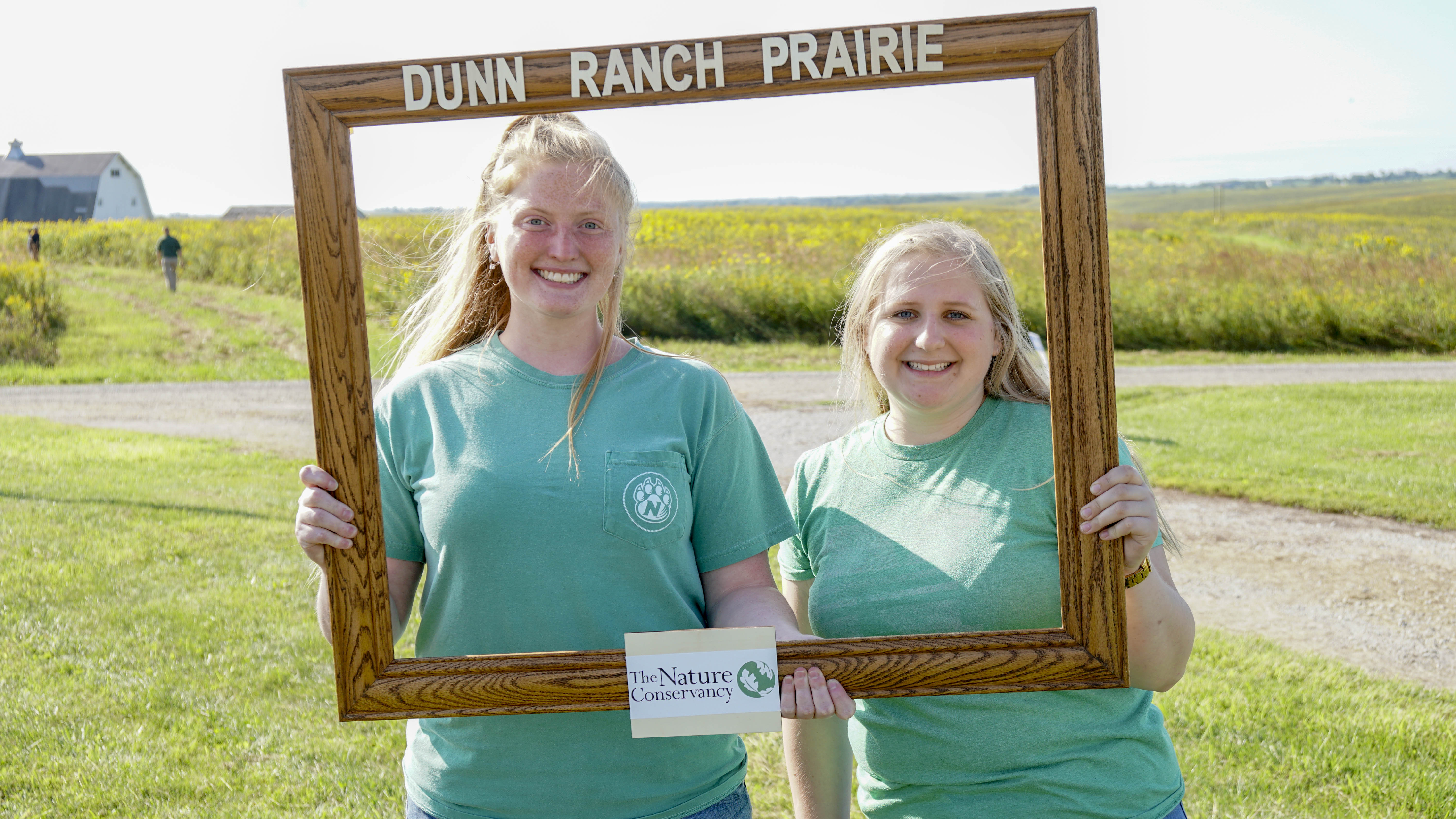 Two women smile and pose while holding up a wooden frame labeled 'Dunn Ranch Prairie.'
