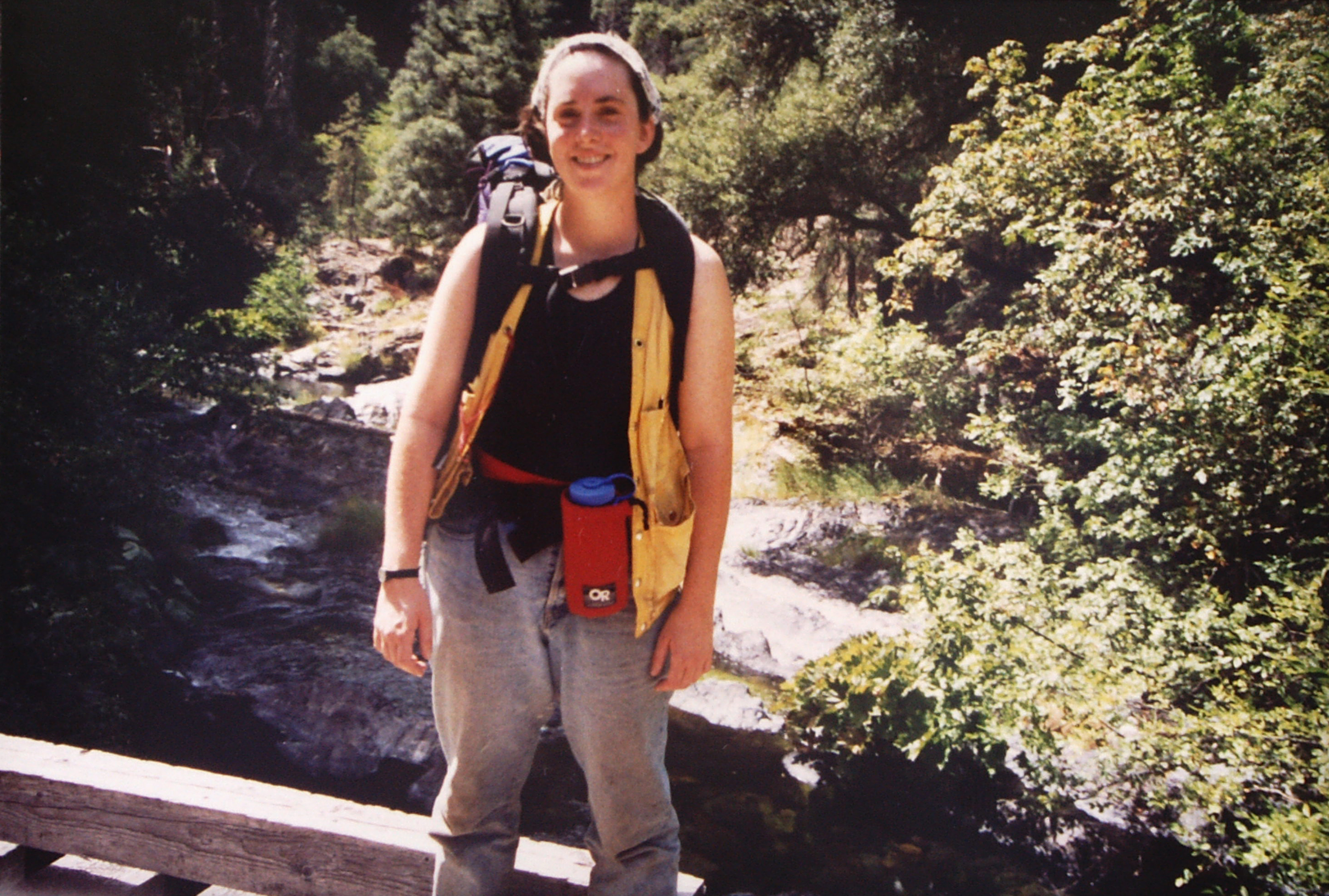 A young Samantha Horn in hiking gear stands smiling on a bridge over a small stream.