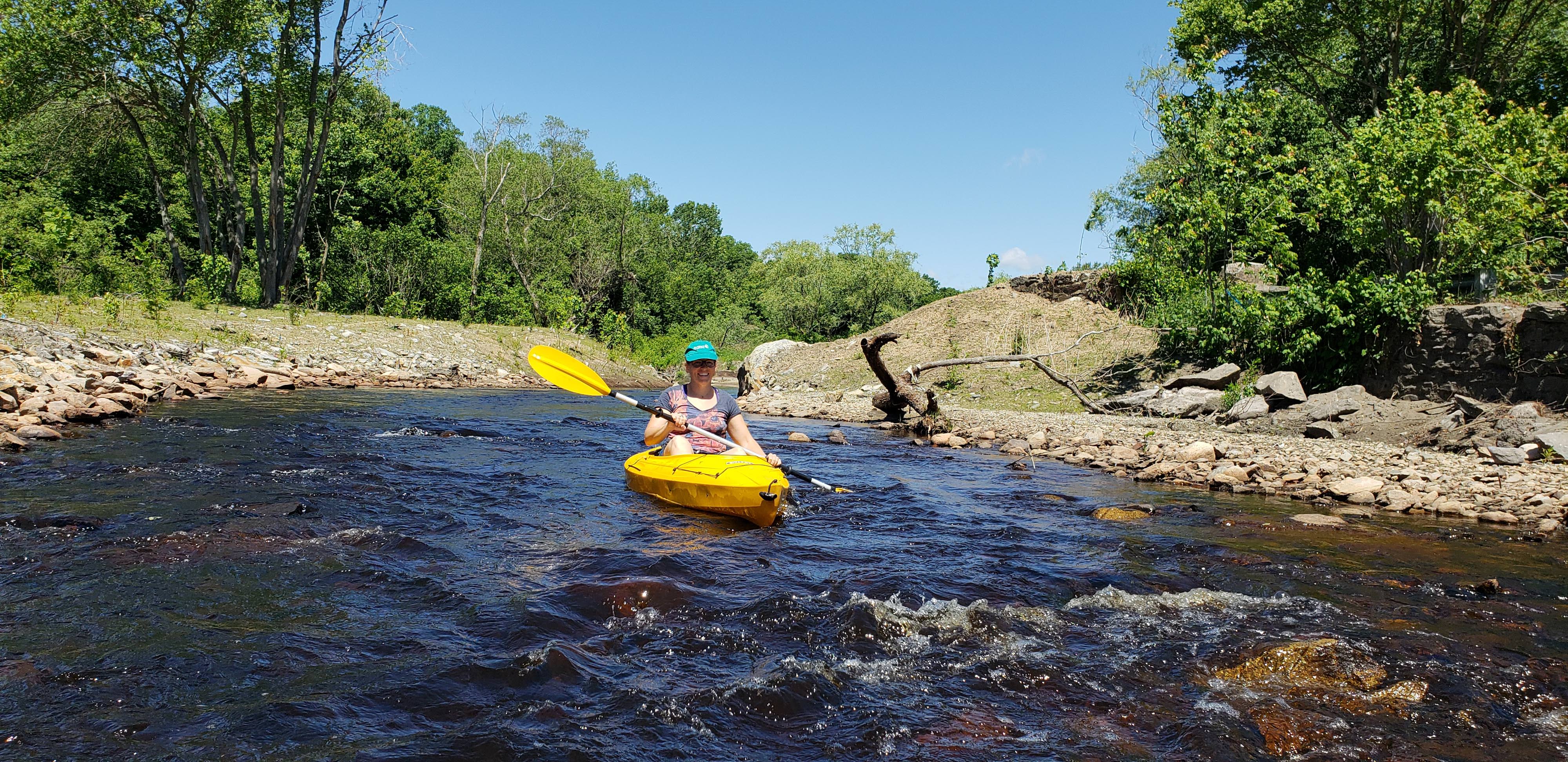 A woman kayaks toward the camera on the Mill River, which is surrounded by rocks and greenery.