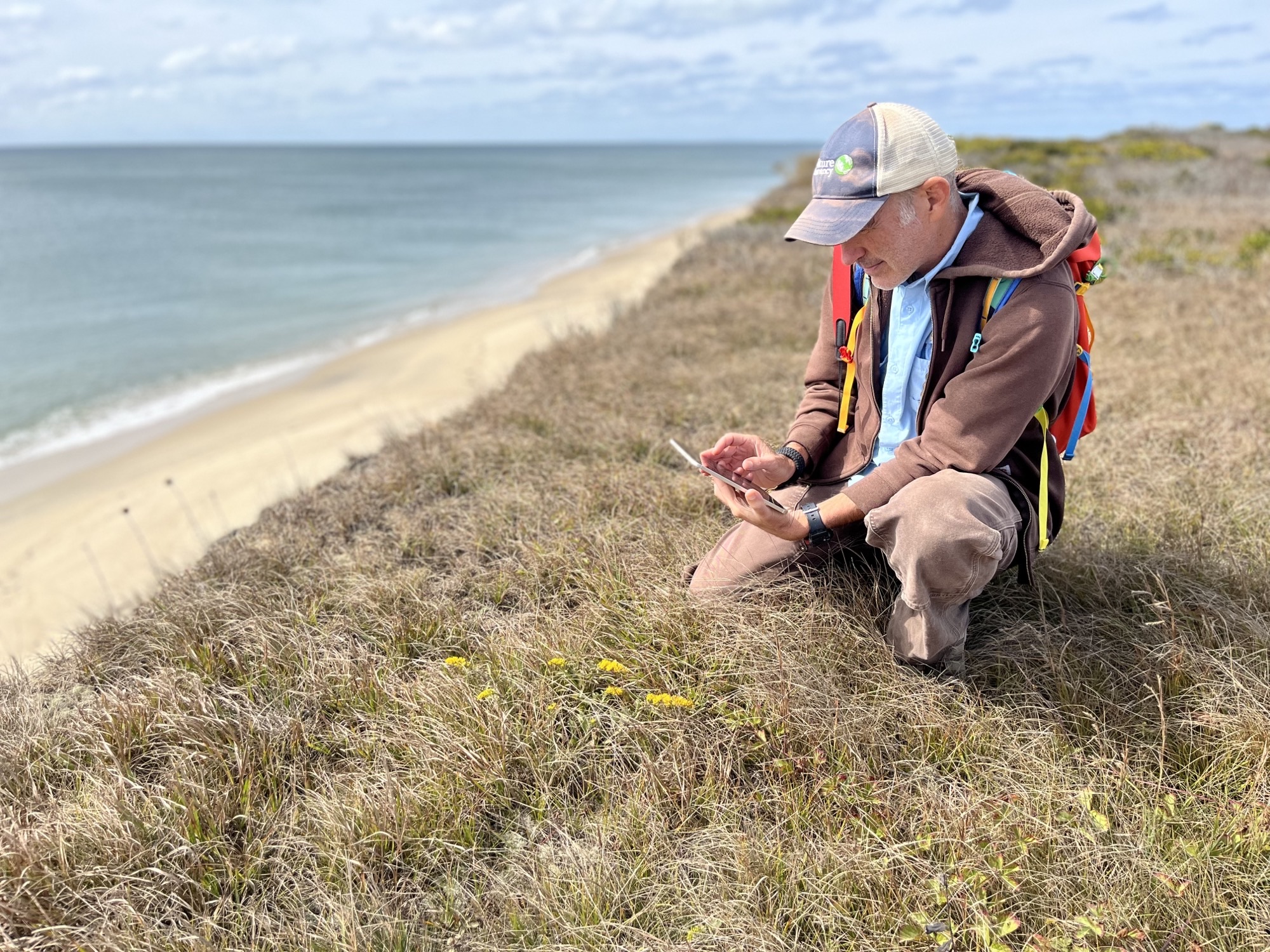 Rich Couse, a seasonal stewardship staff member, kneels on a grassy dune along a beach. 