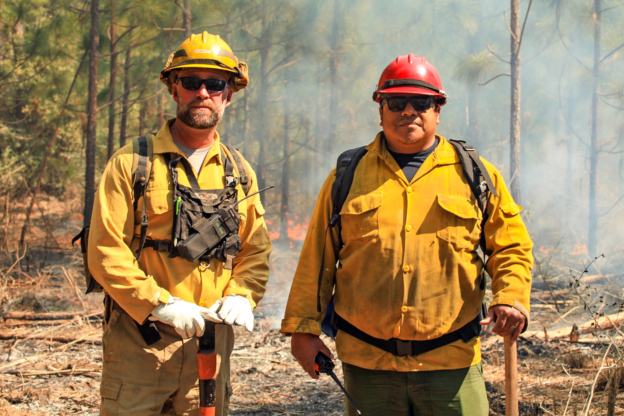 Two men in yellow firefighting gear stand next to each other with axes.