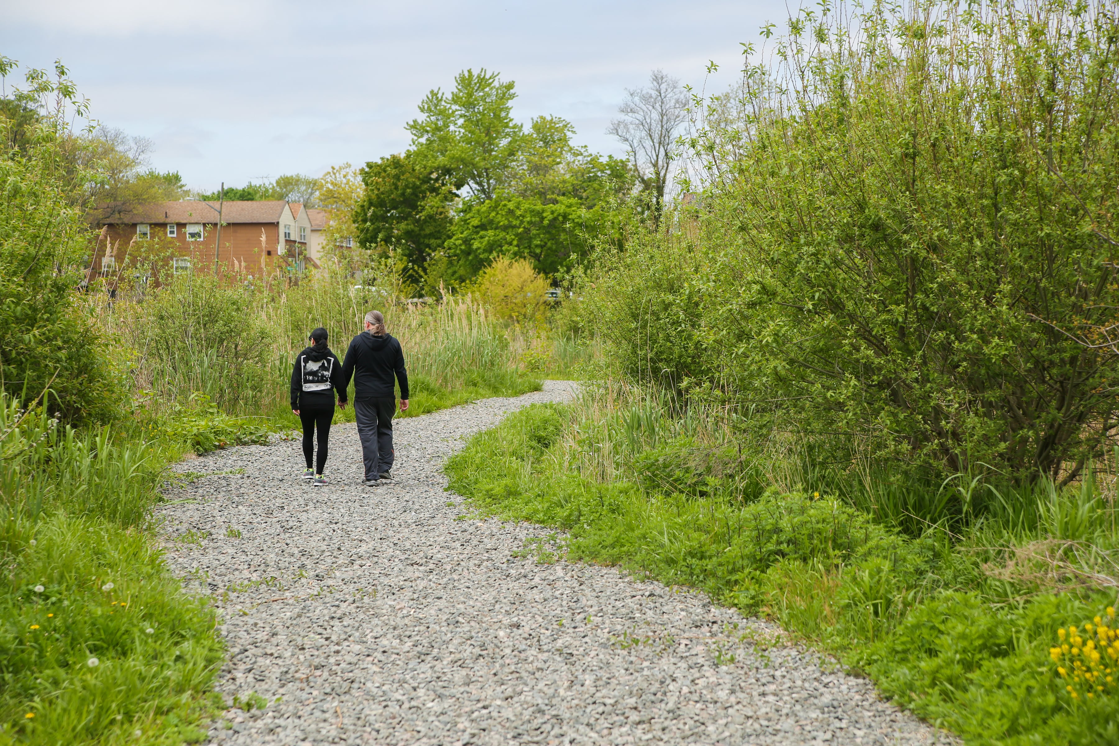 Couple, holding hands, walks on a gravel path surrounded by greenery, with a brick house in the background.