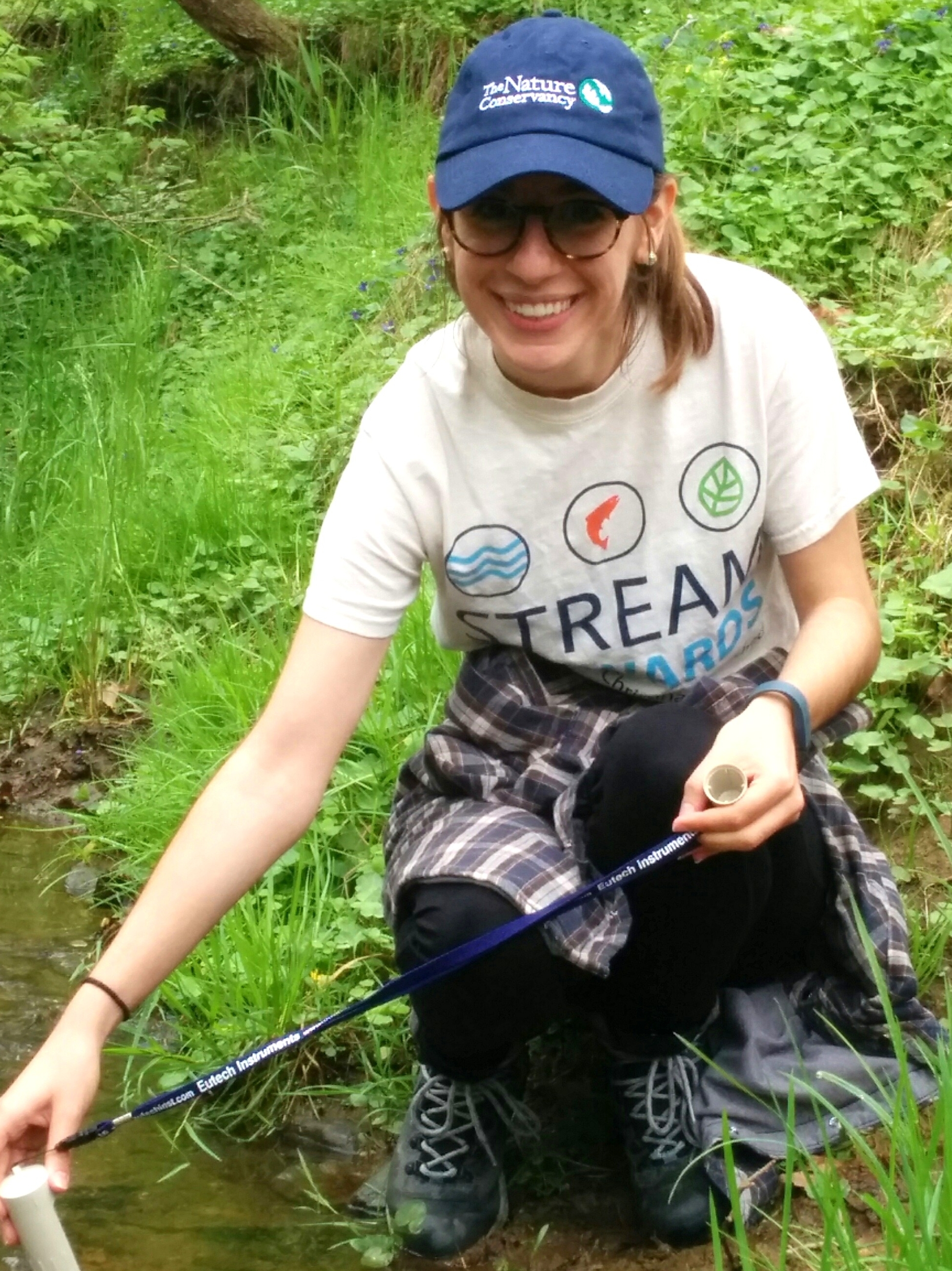 A smiling woman crouches down at the edge of a stream. She is holding one end of a white tube used to sample water quality.