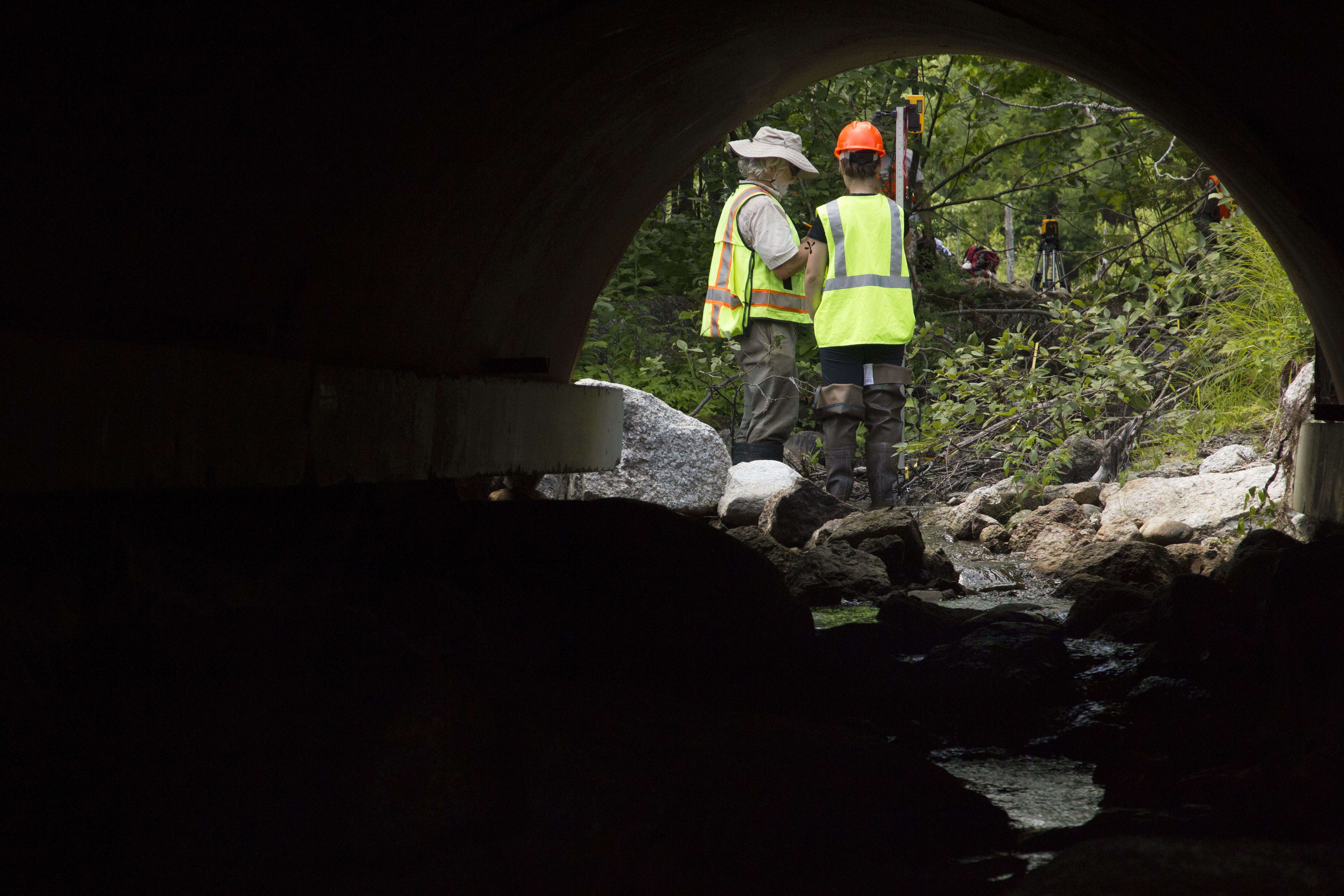 Two construction workers viewed from inside a tunnel under a road.