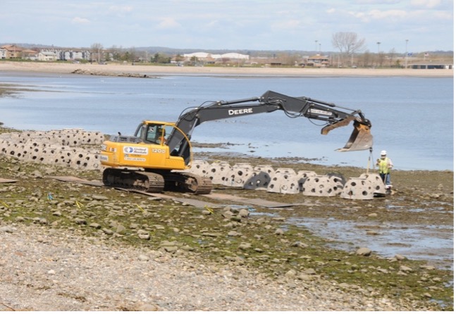 A yellow excavator places concrete reef balls along a shoreline.