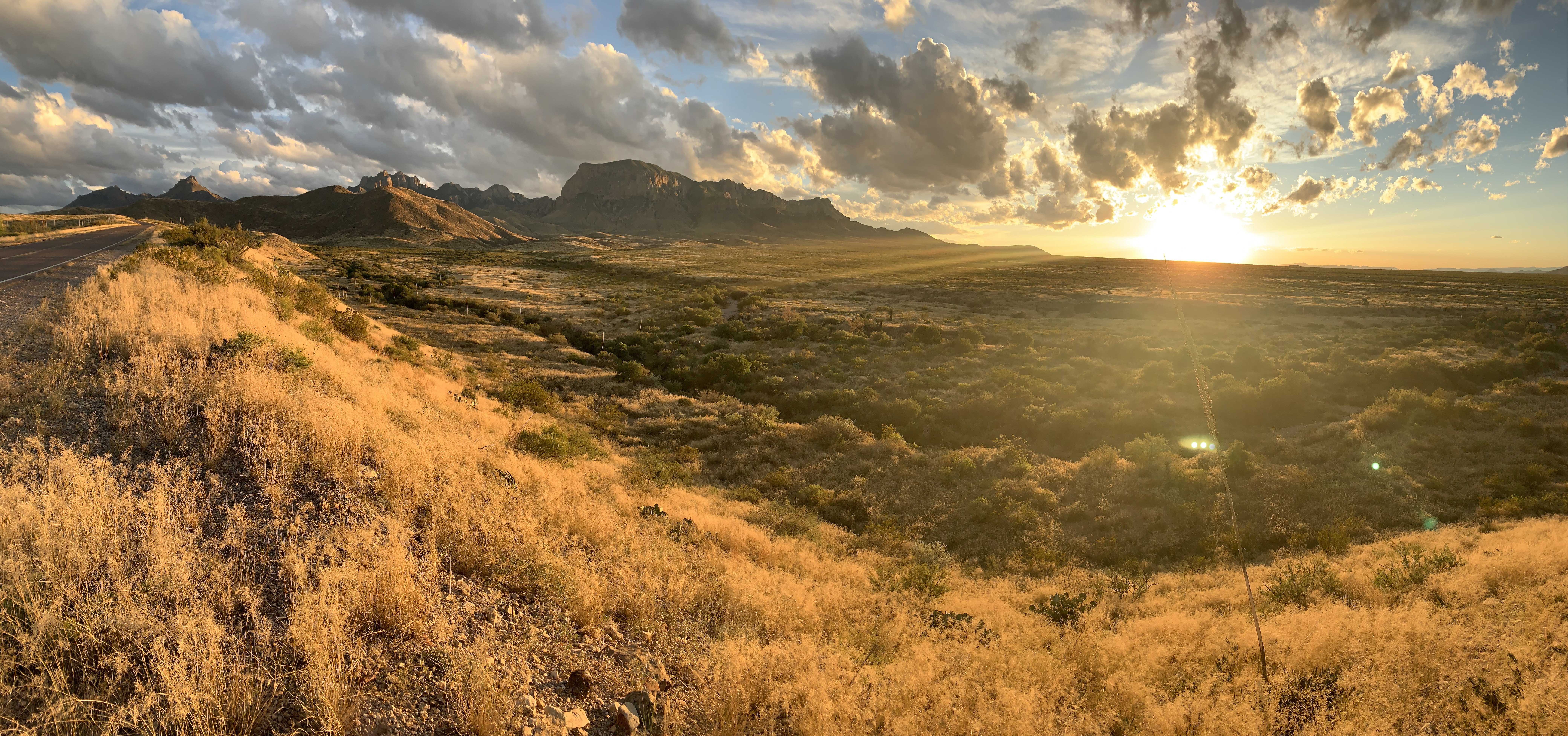 Shrubs and tall grass meet towering rocky mountains as the sun rises along a West Texas road.