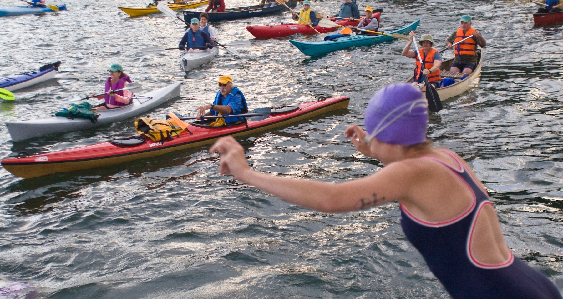A person on the right hand side of the frame is mid-jumping into the water. They are wearing a purple swim cap, goggles, and a one-piece swimsuit. Many kayakers are onlooking.